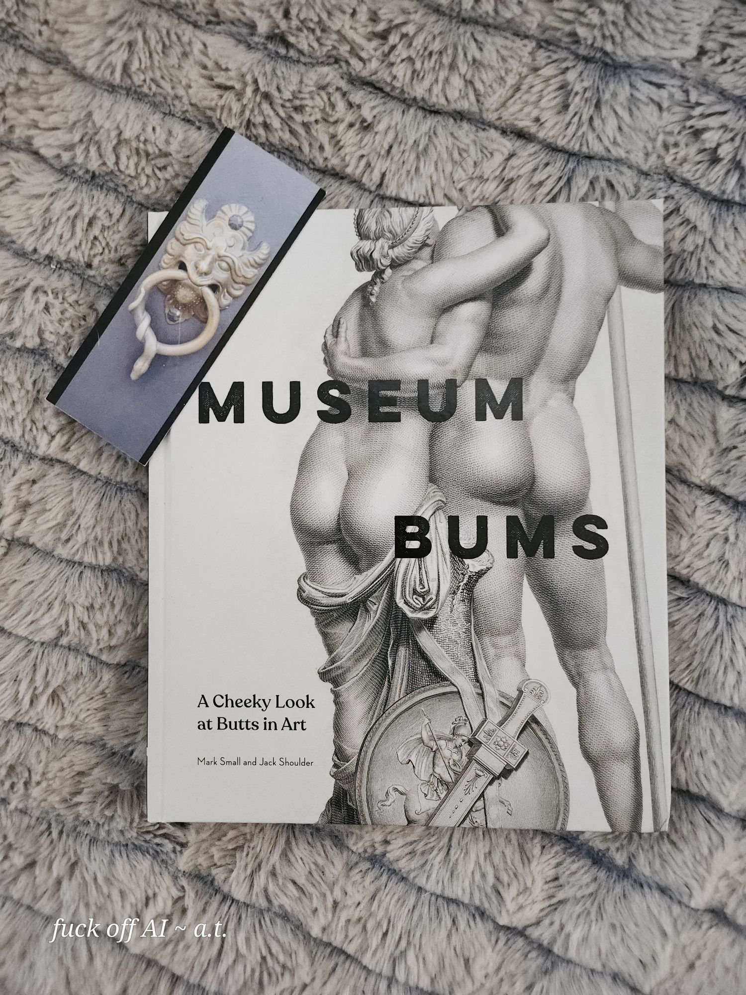 Photo of a white hardcover book sitting on a fluffy gray blanket. Resting on the book's corner is a bookmark with the image of a tan lion-faced door knocker with a ring in its mouth that has a snake curled around it. The book title in large black blocky font: MUSEUM BUMS. The title is laid out over the backside of a statue (possibly Greek or Roman) of a naked man and woman holding each other. A cloth wrap has mostly fallen off the woman and is wrapped loosely around her legs. At the base is a round shield and a short sword in a decorative sheath. Image of the statue is in black and white.  Smaller subtitle on lower left in black font: A Cheeky Look at Butts in Art. In tiny font underneath: Mark Small and Jack Shoulder