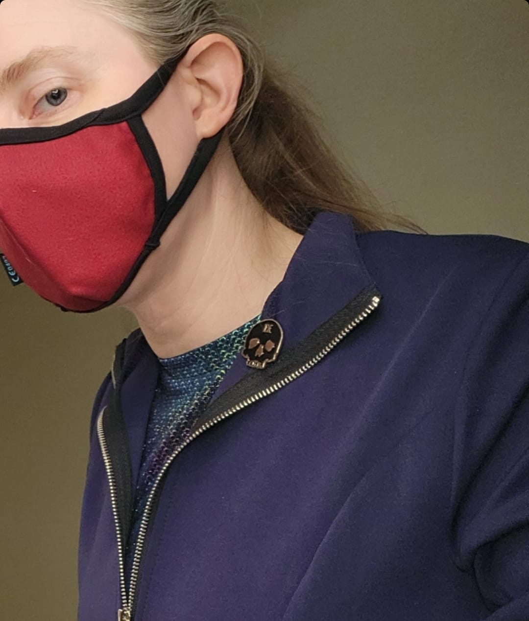 Photo: Close-up of pale caucasian woman from the chest up and head cocked at an angle with the top cropped off, creating one-eyed peekaboo effect. Dark blond hair pulled back. Wearing red fabric filter mask. Eyes are blue. Dark blue-purple fabric thin coat unzipped, but only upper chest and shoulders shown. There is a multi-hued patterned shirt underneath (shiny like a mermaid's fins). Close-up up of pin on lapel of a black skull with a golden IX on its forehead. Tan hotel room wall in background.