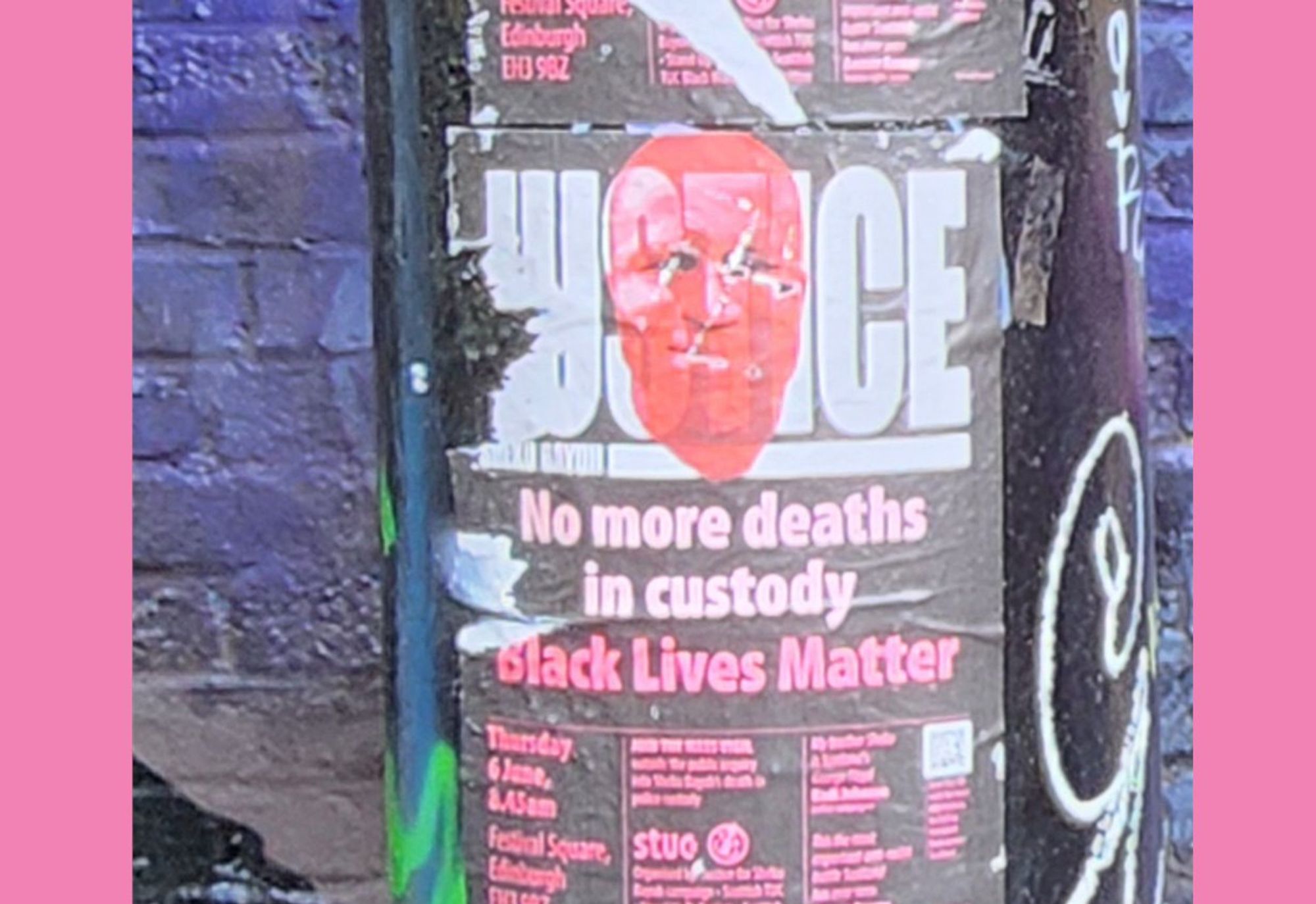 Photo of black pole with flyers and grafitti on it. In white and pink text on the black flyer: JUSTICE (red face overlaid on justice) / No more deaths / in  custody / Black Lives Matter / (Meeting details and other text too small to decipher.) There is a purple spray-painted brick wall behind the pole.