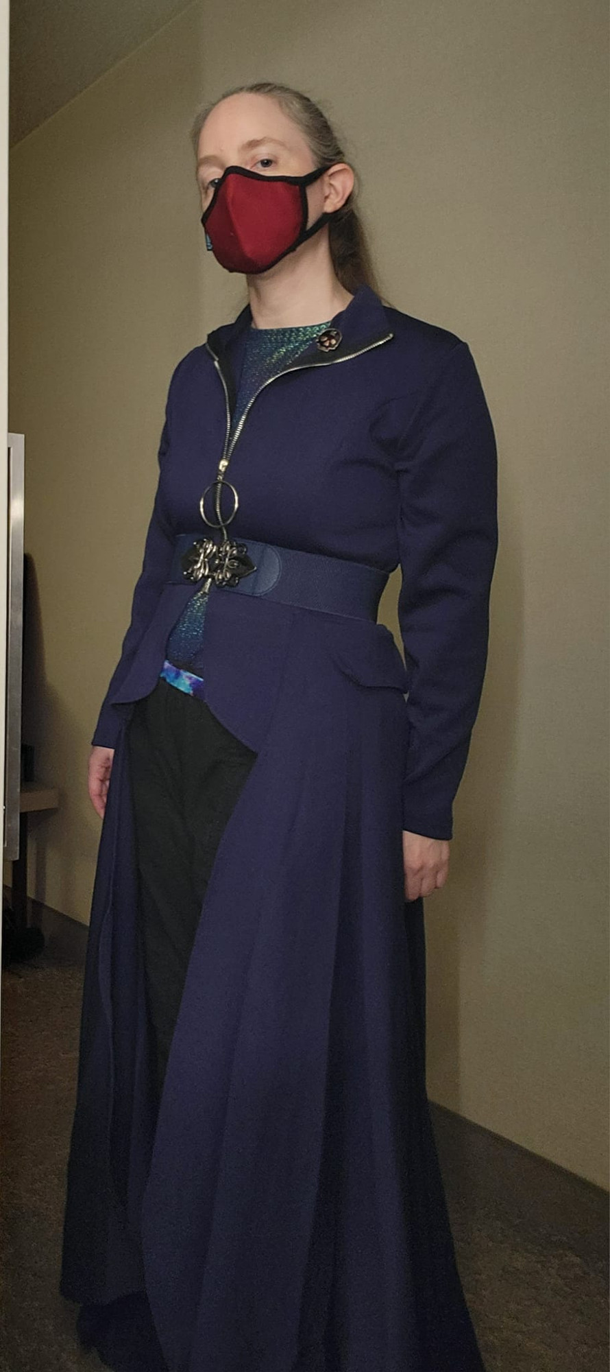 Pale caucasian woman with dark blonde hair pulled back wearing red fabric filter mask. Dark blue-purple fabric thin coat unzipped down to high waist belt with gold clip. Long sweeping coat tails that curves open at the waist. Black pants underneath. Hotel room background tan wall, brown carpet.