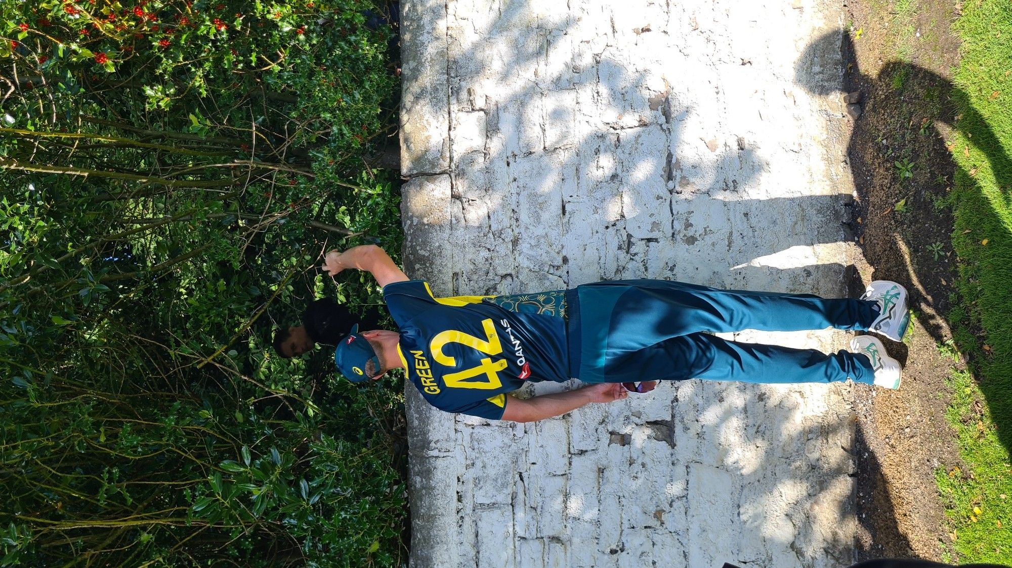 Australian cricket player in green playing kit facing away from camera reaching over stone wall with right hand to get ball back, jersey says Green with numerous 42 on back, grass and dirt at bottom of shot, dark green foliage above and behind wall