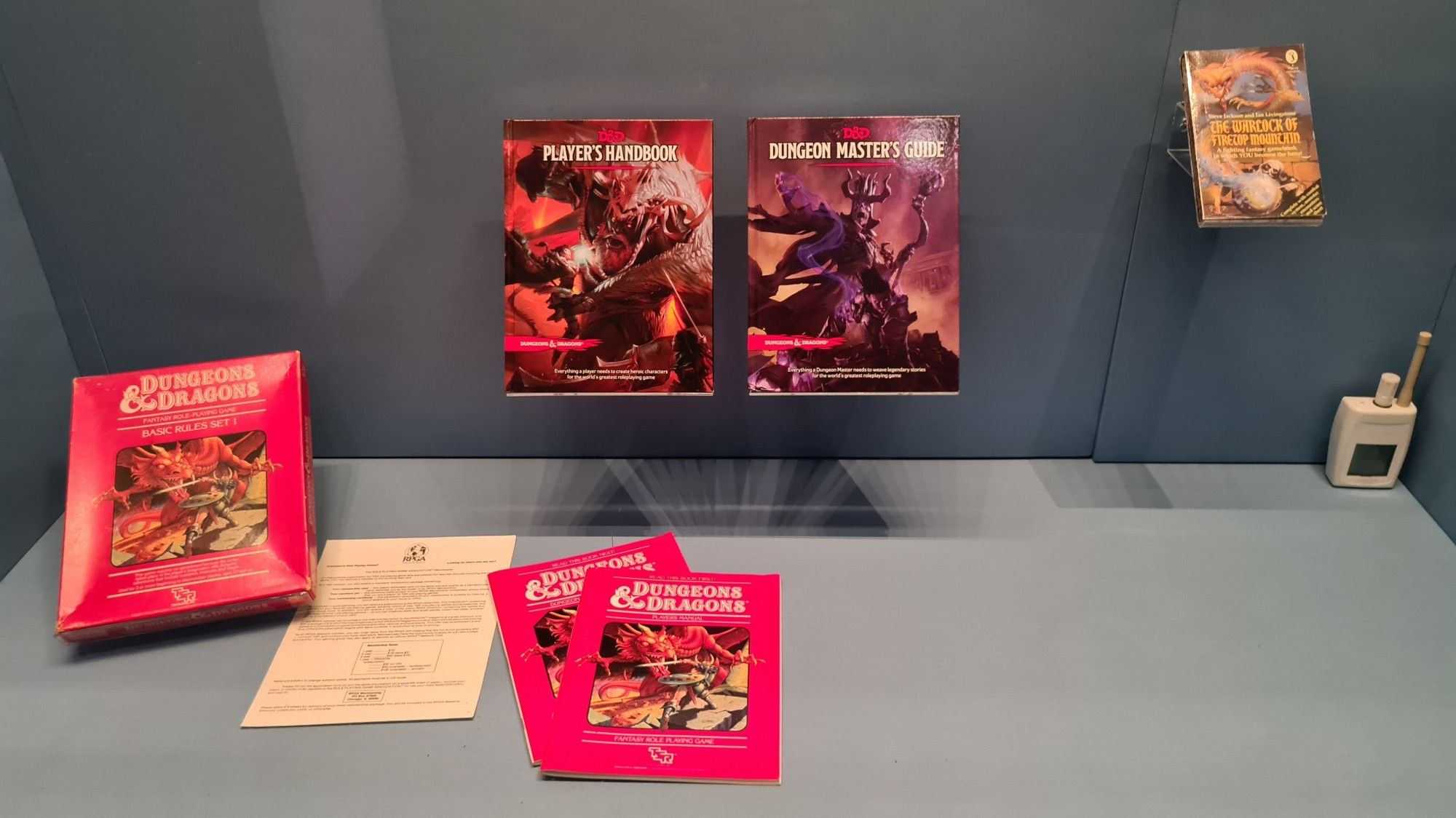 Display cabinet housing role playing games and books,  steel blue background, left of shot is Dungeons and Dragons basic red box set with the rulebooks spread our next to it, centre is the current 5th edition D and D Players Handbook and Dungeon Masters Guide, to the right an original copy of the Fighting Fantasy 1st book The Warlock of Firetop Mountain