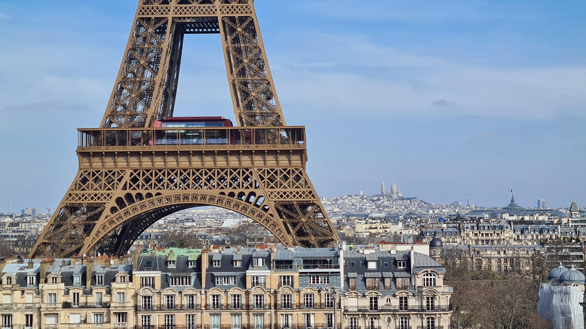 Eiffel tower close up have middle third approx. centre of shot, blue sky with whisky blue clouds frames the tower,  Parisian apartments below the tower behind the tower the Paris skyline stretches away with hill of Montmartre and the towers of the Sacre Coeur Basilica just visible in the distance