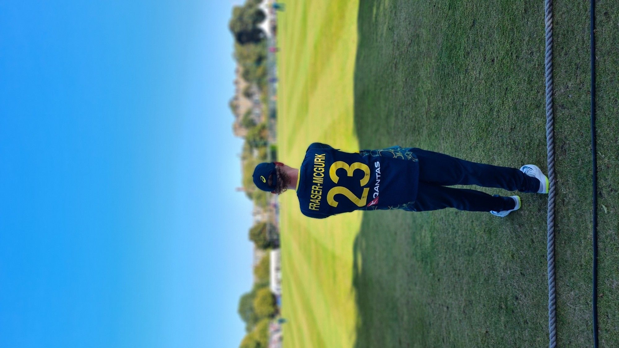 Australian cricket player in green playing kit facing away from camera standing on boundary, jersey says FraserMcGurk with number 23 on back, out of focus behind player is Grange cricket ground in Edinburgh bathed in sunshine,  Blue sky overhead with light line of clouds, the old grandstand and buildings back middle ground and the green ground bottom of shot with various players in view