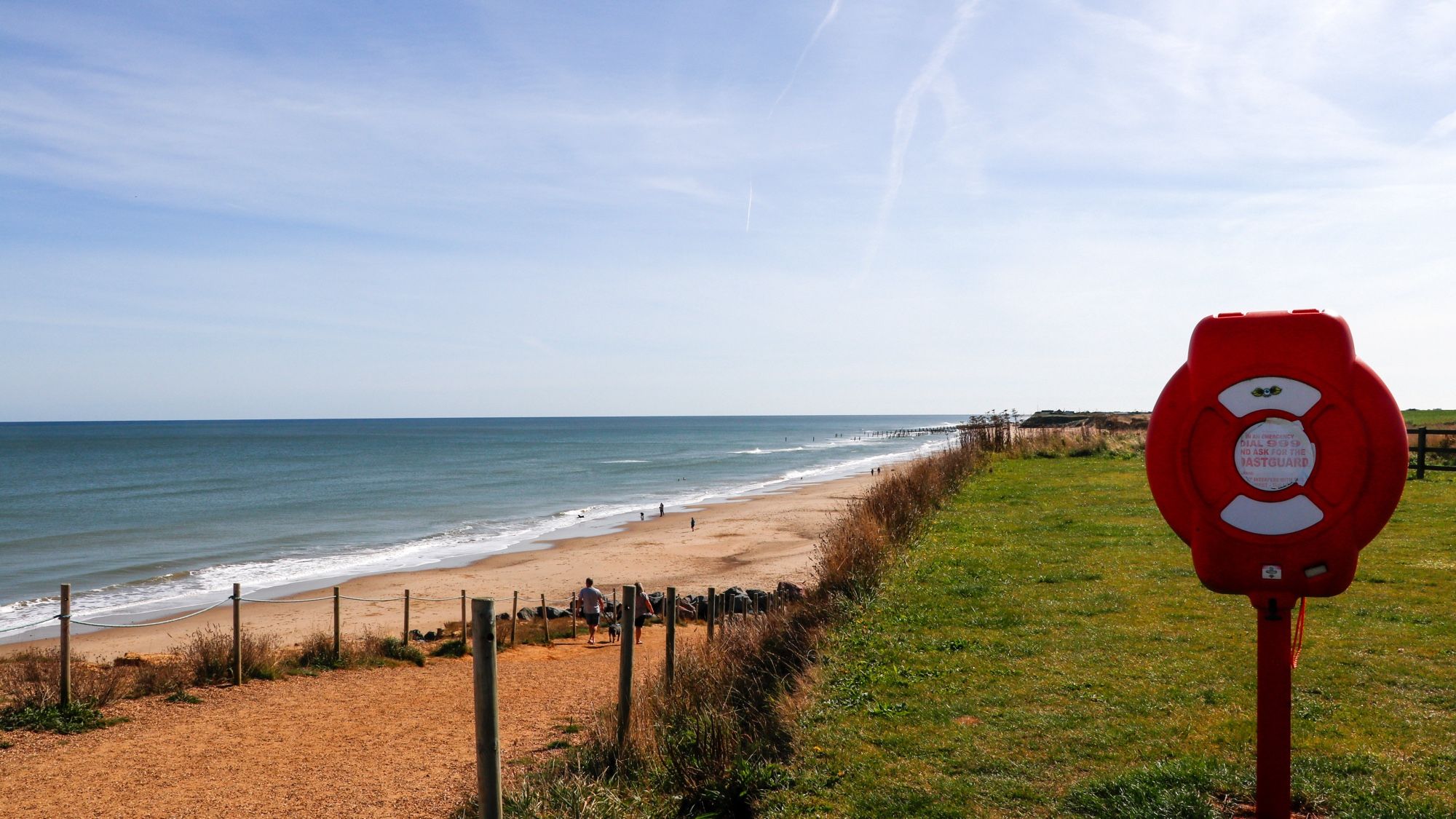 The view from the beach slipway. Hazy blue sky, bright blue sea and yellow sand to the left and green grass on the cliff to the right. In the foreground is a bright red life ring