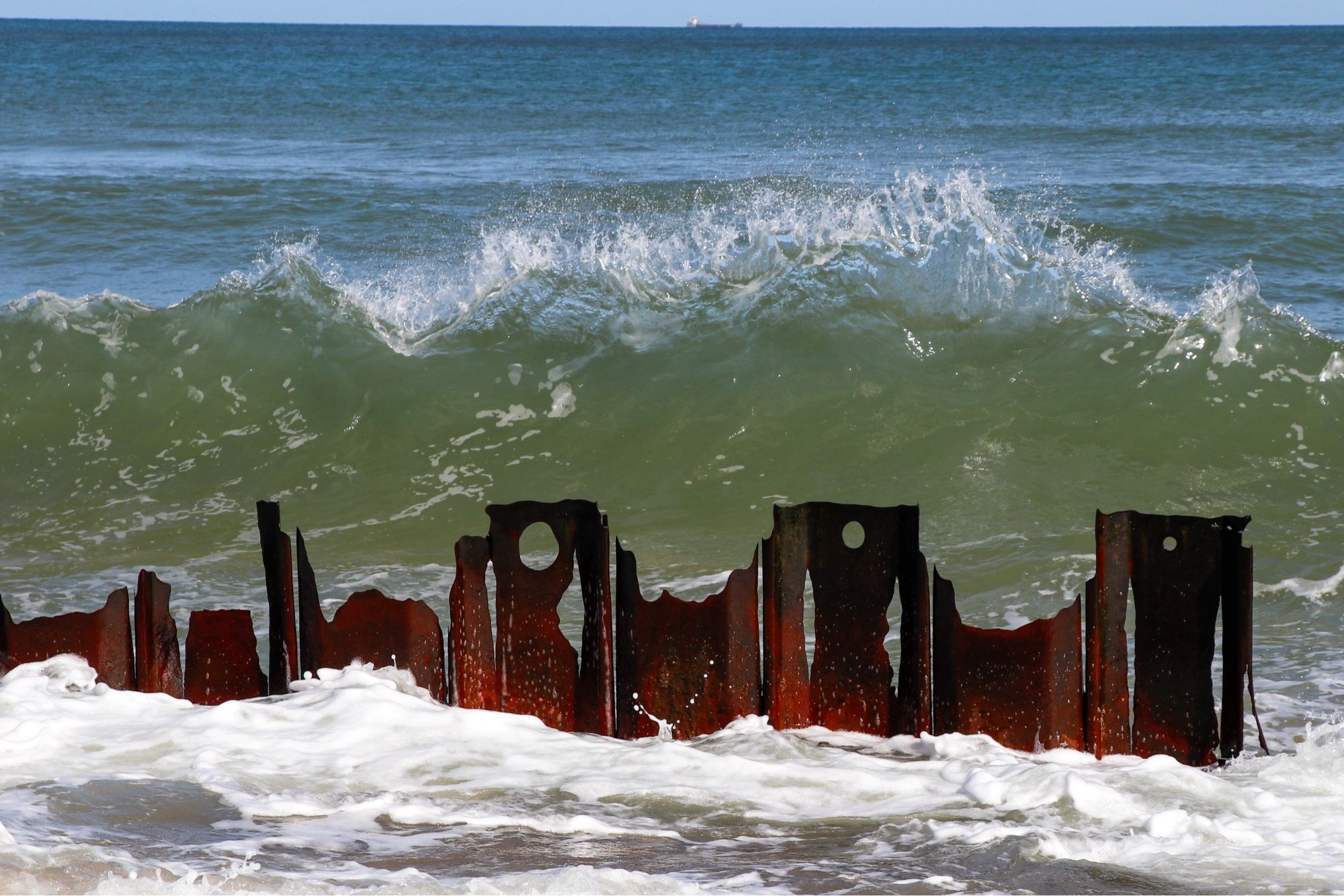 A large white topped green wave behind a red metal beach defence. The white foam from the preceding wave is in the foreground