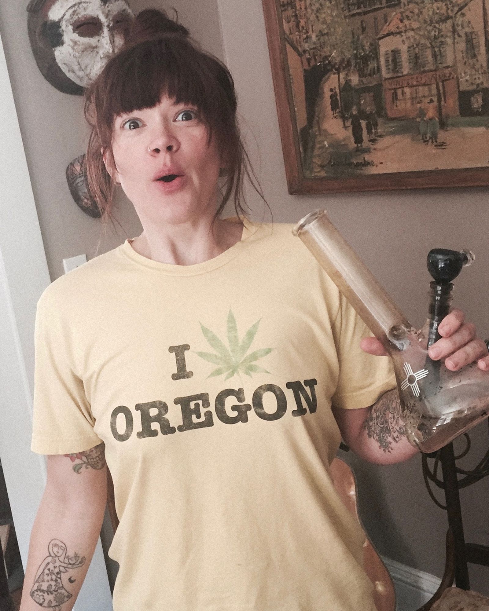 zia mccabe, wearing a faded yellow "i love oregon" t-shirt with the heart replaced with a pot leaf, is holding a glass bong with a zia symbol on it.