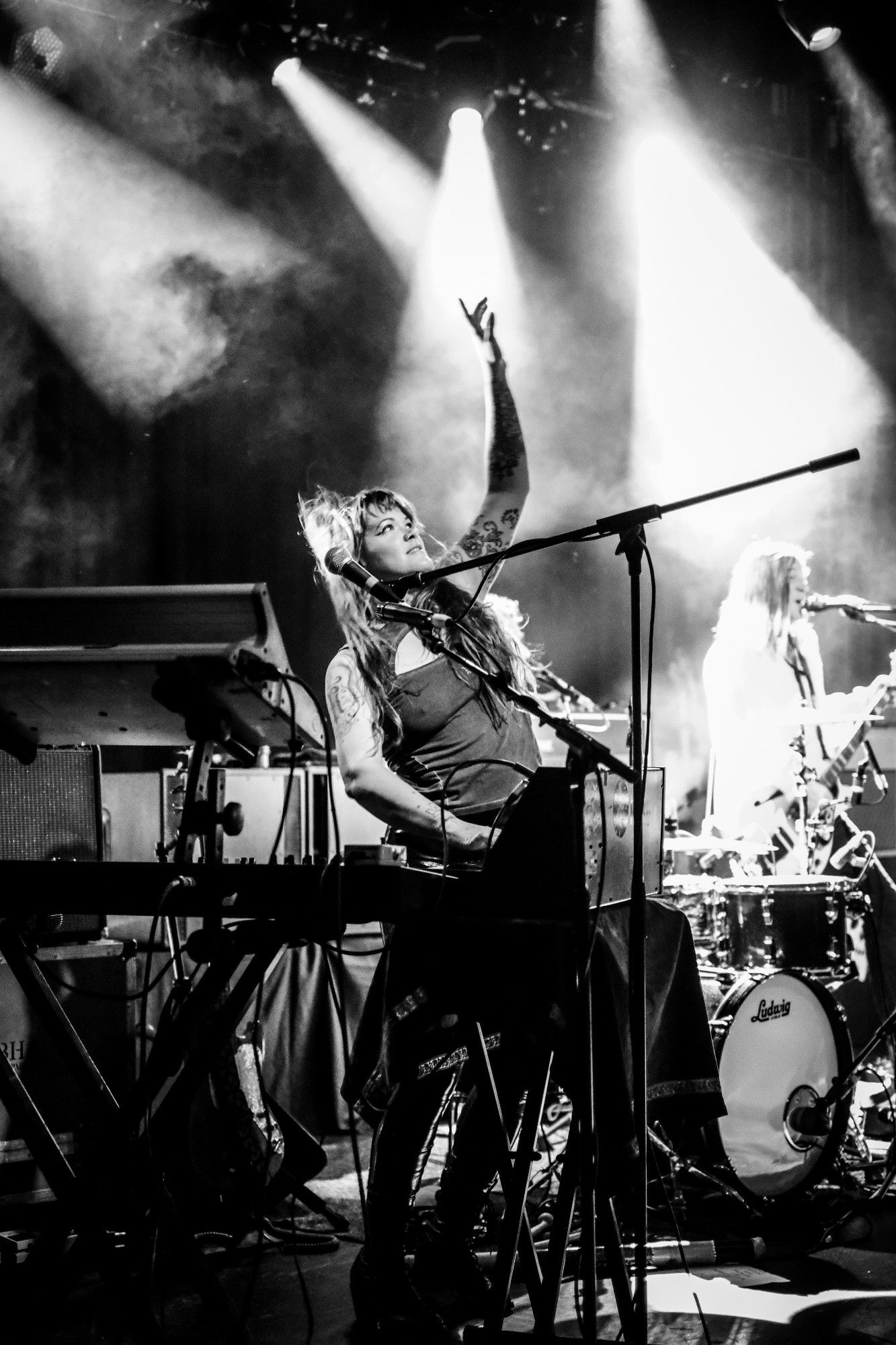 Black and white photo of Zia on stage behind her keyboards pointing up into the spotlit sky.