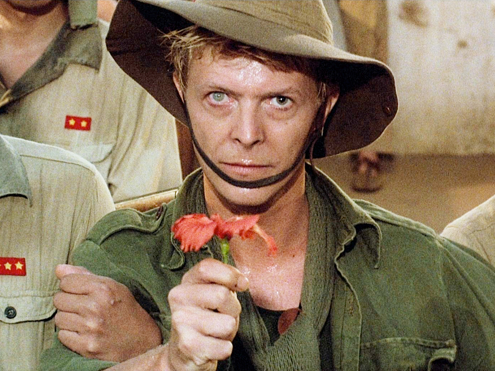 David Bowie as Major Jack Celliers in Merry Christmas, Mr Lawrence, wearing green fatigues and holding out a festive red flower