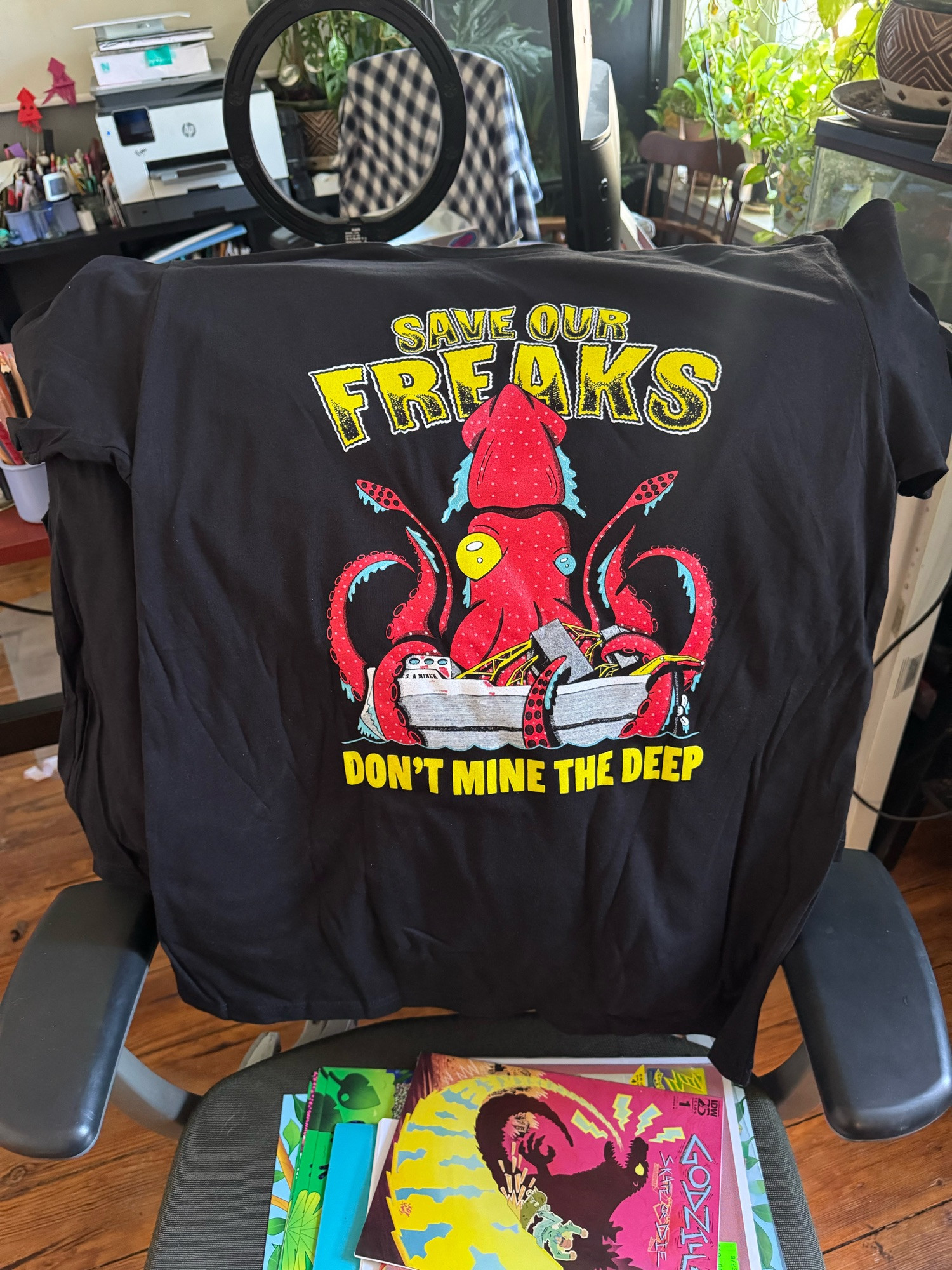 A shirt draped on an office chair with a big ass strawberry squid that’s sinking a mining ship that says in yellow font “save our freaks don’t mine the deep”