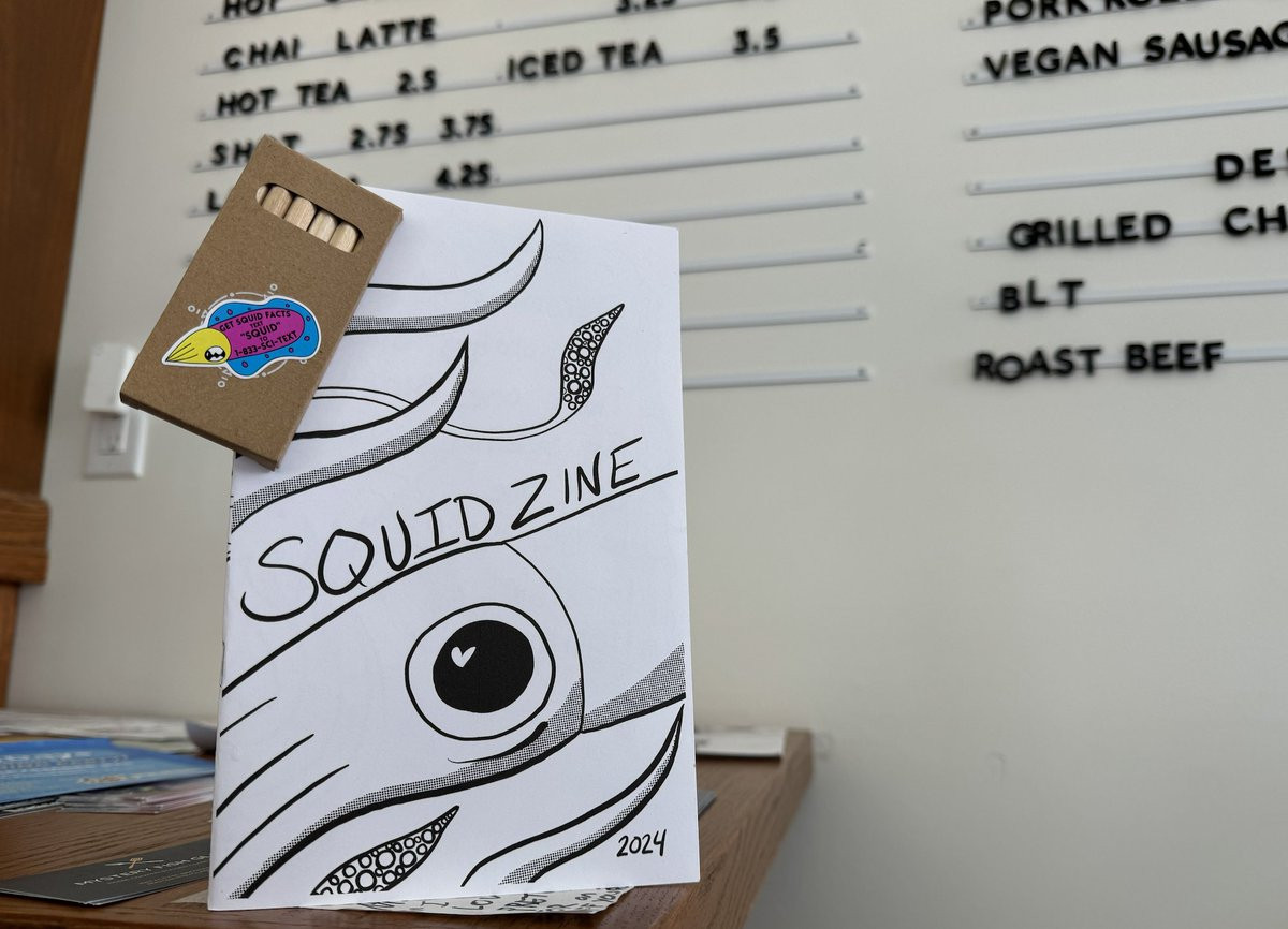 Squid zine with a pack of colored pencils paperclipped to the corner, on a coffee bar with a coffee menu in the background. The zine is black and white with a bit squid on it, very zoomed it and it says Squid Zine