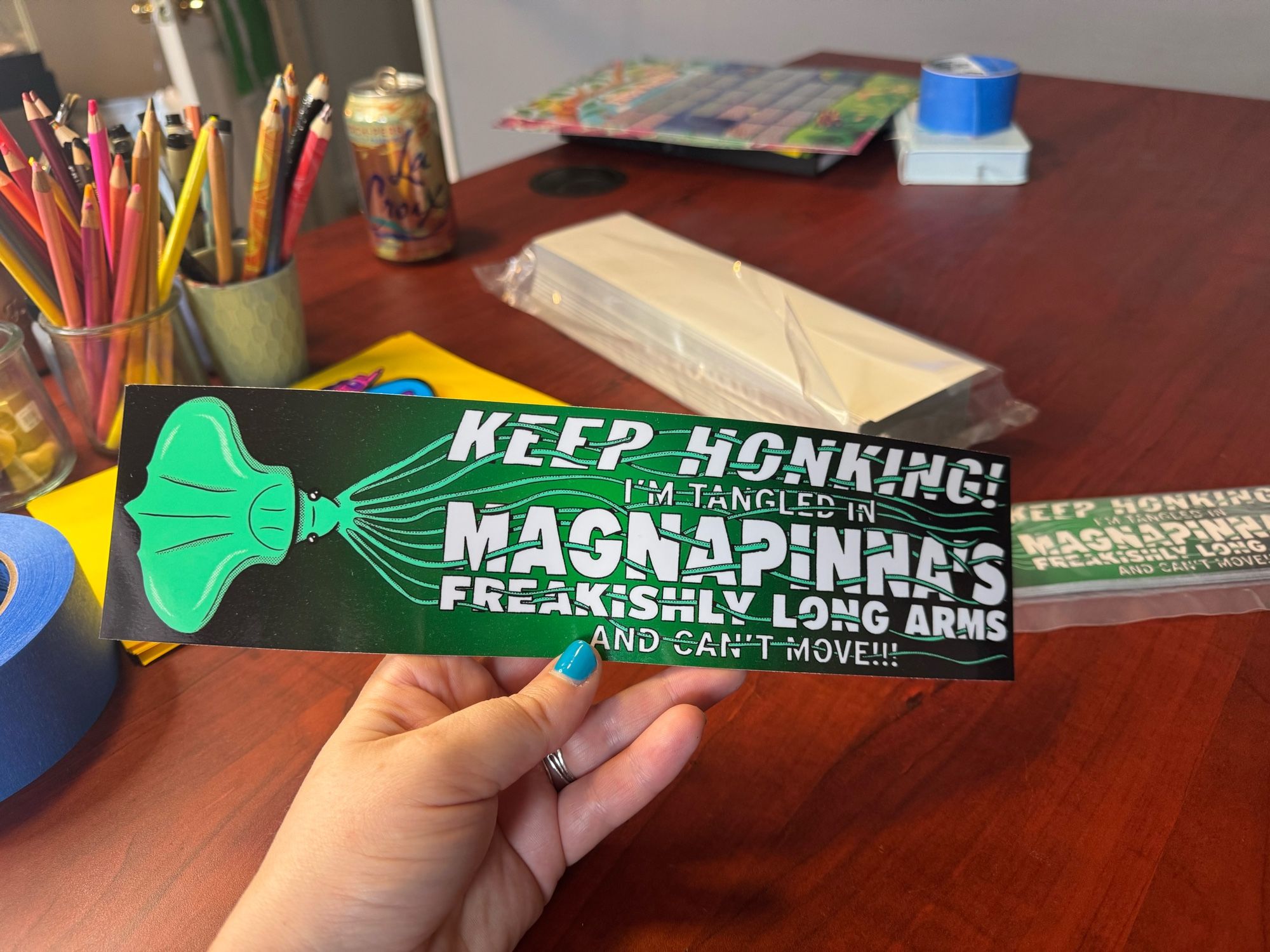 Sarah holds up a bumper sticker that says “KEEP HONKING! I’m tangled in magnapinna’s freakishly long arms and can’t move!!!!” With magnapinna’s arms all winding through the letters