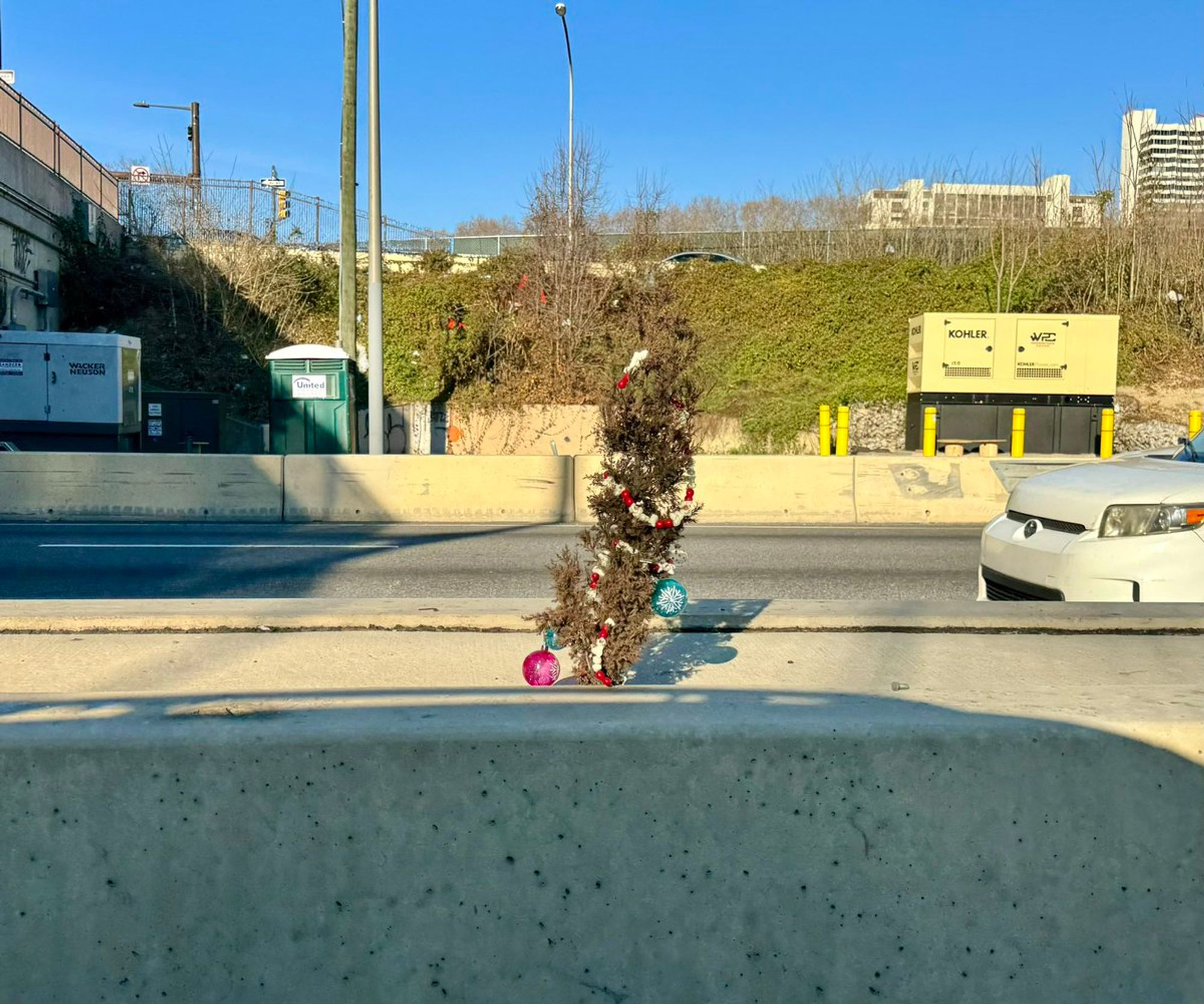 A 1.5-2 foot tall little tree with red and white garland and Christmas ornaments growing out of the divider of a highway