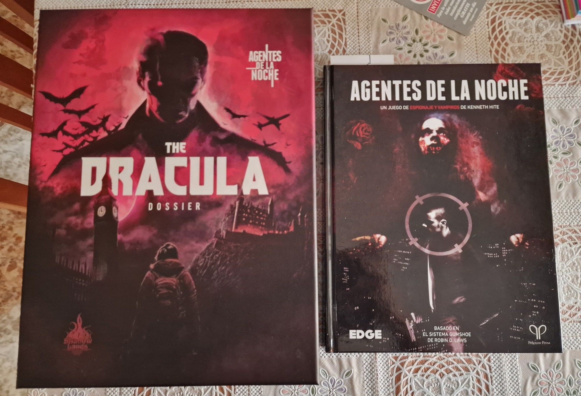 Caja de la campaña The Dracula Dossier. La caja tiene diferentes tonalidades rojizas con Drácula en la parte superior donde le sobrevuelan unos murciélagos en el lado izquierdo y un avión en el lado derecho. En primer plano vemos a una mujer vestida con un abrigo y portando una mochila. Entre ambos se ven un reloj que podría ser el Big ven y un castillo sobre una una colonia. 

A la derecha de la caja en la foto está el manual básico de agentes de la noche.