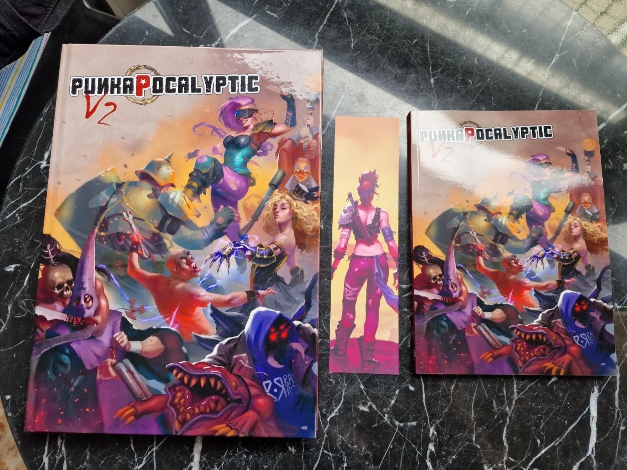 Reglamento tamaño A4 tapa dura de Punkapocalyptic, marcapáginas con Lizzy de espalda y reglamento de bolsillo de Punkapocalyptic (A5). La portada es una escaramuza con representantes de todas las facciones del juego