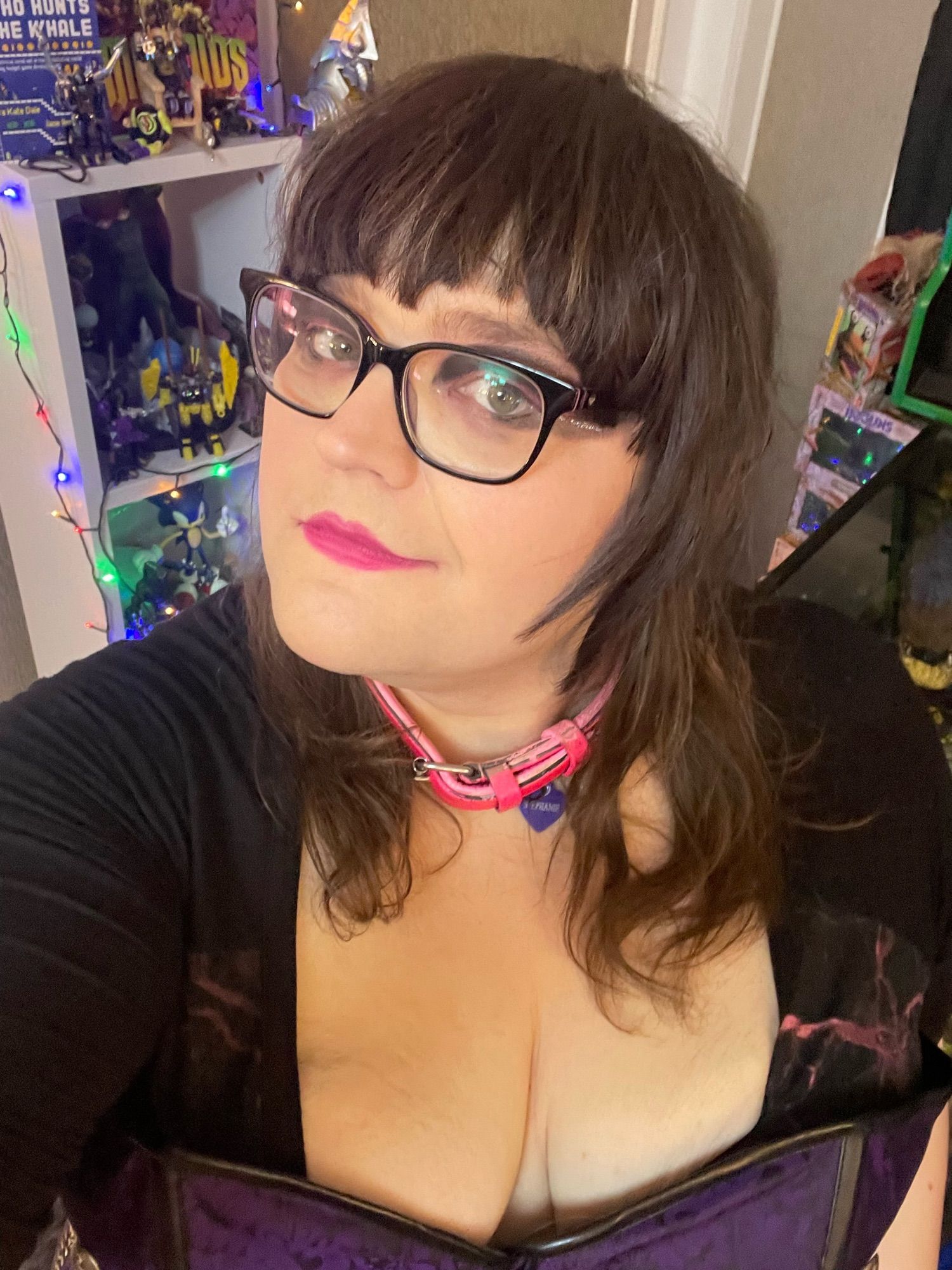 Stephanie Sterling selfie - pink lipstick, black/silver eyeshadow, with black glasses and pink collar, inc. corset-assisted cleavage.