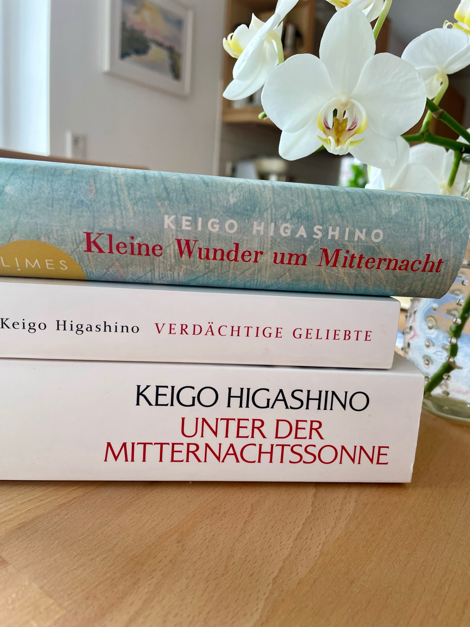 Zu sehen ist ein Stapel von drei unterschiedlichen Büchern auf einem Holztisch, im Hintergrund die Blüten einer weißen Orchidee