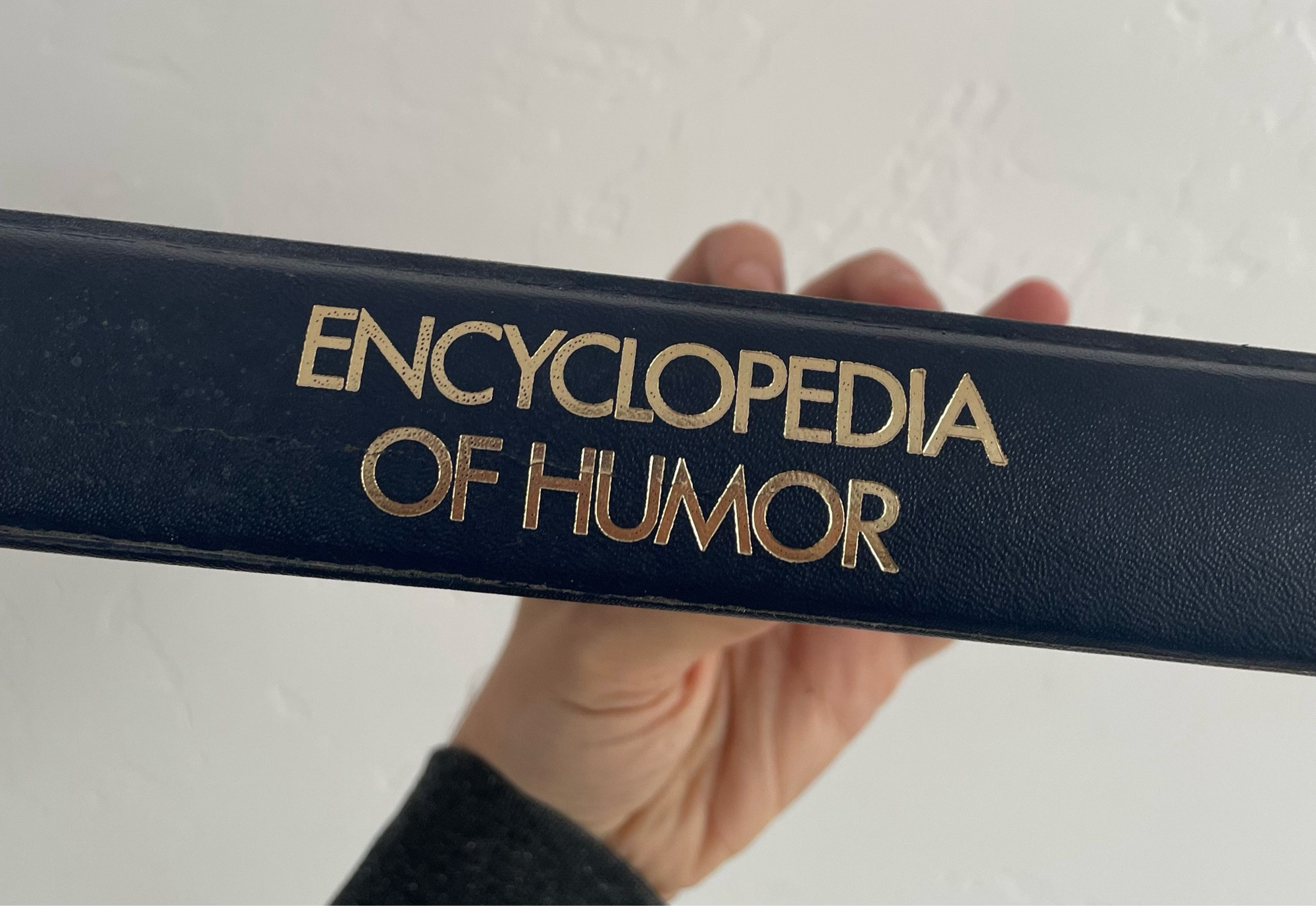 My hand holding a book called ENCYCLOPEDIA OF HUMOR, spine facing the camera with the title printed on it in gold