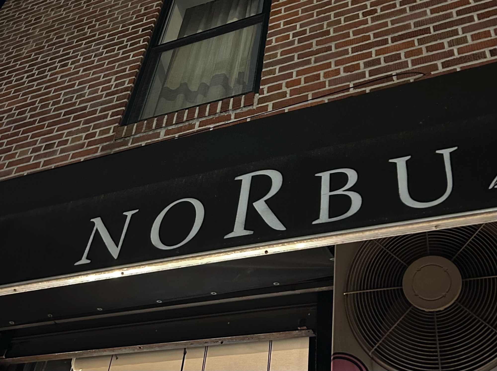 NORBU

(Written on Black awning in white text beneath a brick building wall with a window in it, above a fan)