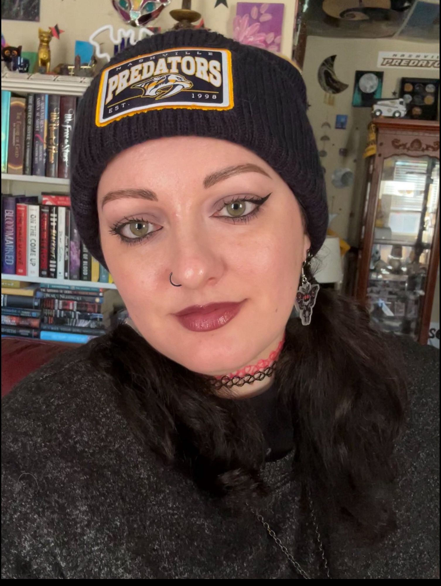 Selfie of me in a Preds beanie, black winged eyeliner, and purple ish lip color. I was wearing a gray long sleeved shirt and mothman earrings. Also 2 of those stretchy tattoo chokers, one black and one hot pink.