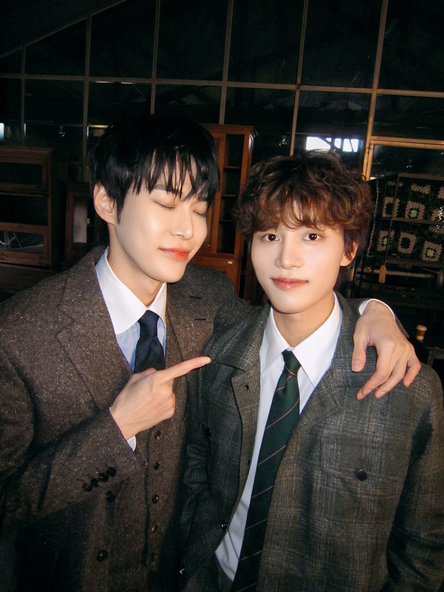 Our main vocalists, Doyoung and Taeil. They are wearing their wool suits from the Be There For Me music video. Doyoung has his eyes closed, is smiling, has his arm around Taeil, and is pointing at Taeil.