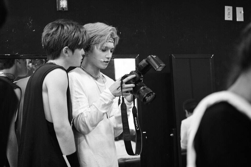 Jaehyun looks over Taeyong’s shoulder as Taeyong looks at a picture he took on the screen of a professional digital camera. The picture is in black and white.