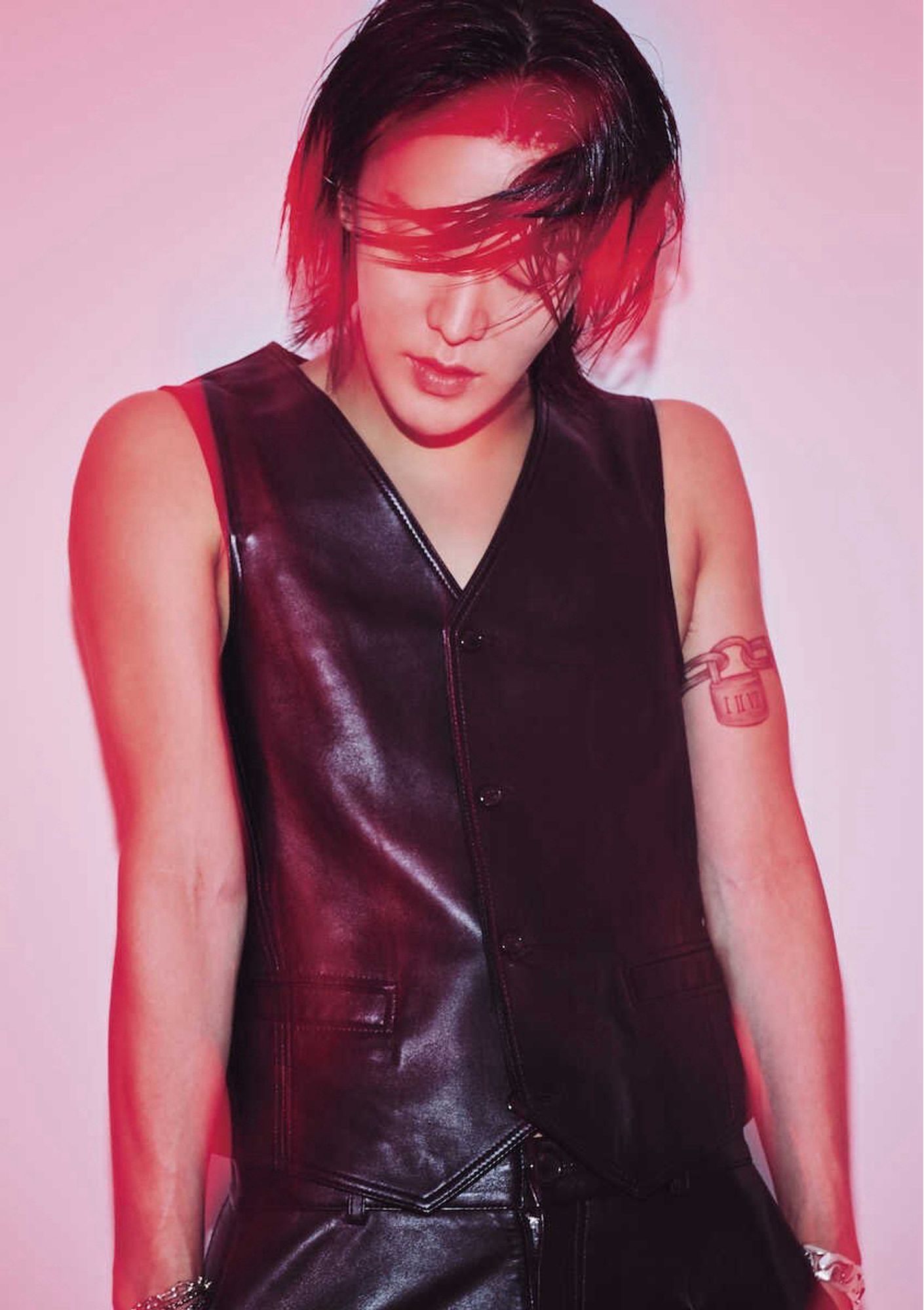 Yuta wearing a leather fest and leather pants, with his hands in his pockets. He’s standing in front of a main wall. His hair is long and wet, with a front section swinging in front of his face covering his eyes. The picture has a red tone to it, as though a red light is being shone on him.