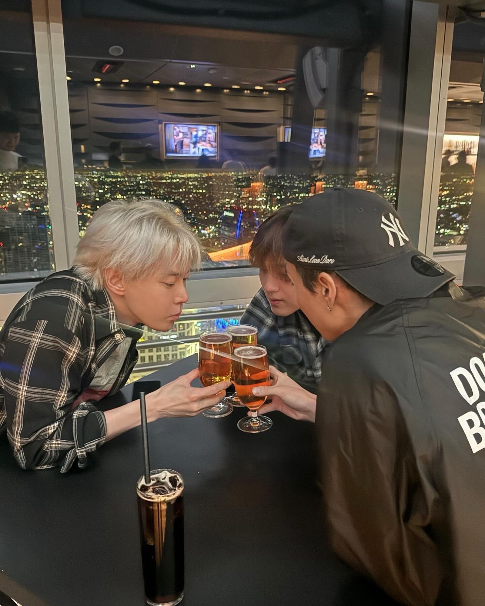 Doyoung, Haechan, and Johnny sitting around a restaurant table, the city lights below them can be seen through the window behind them. They are cheers-ing each with a glass of beer.