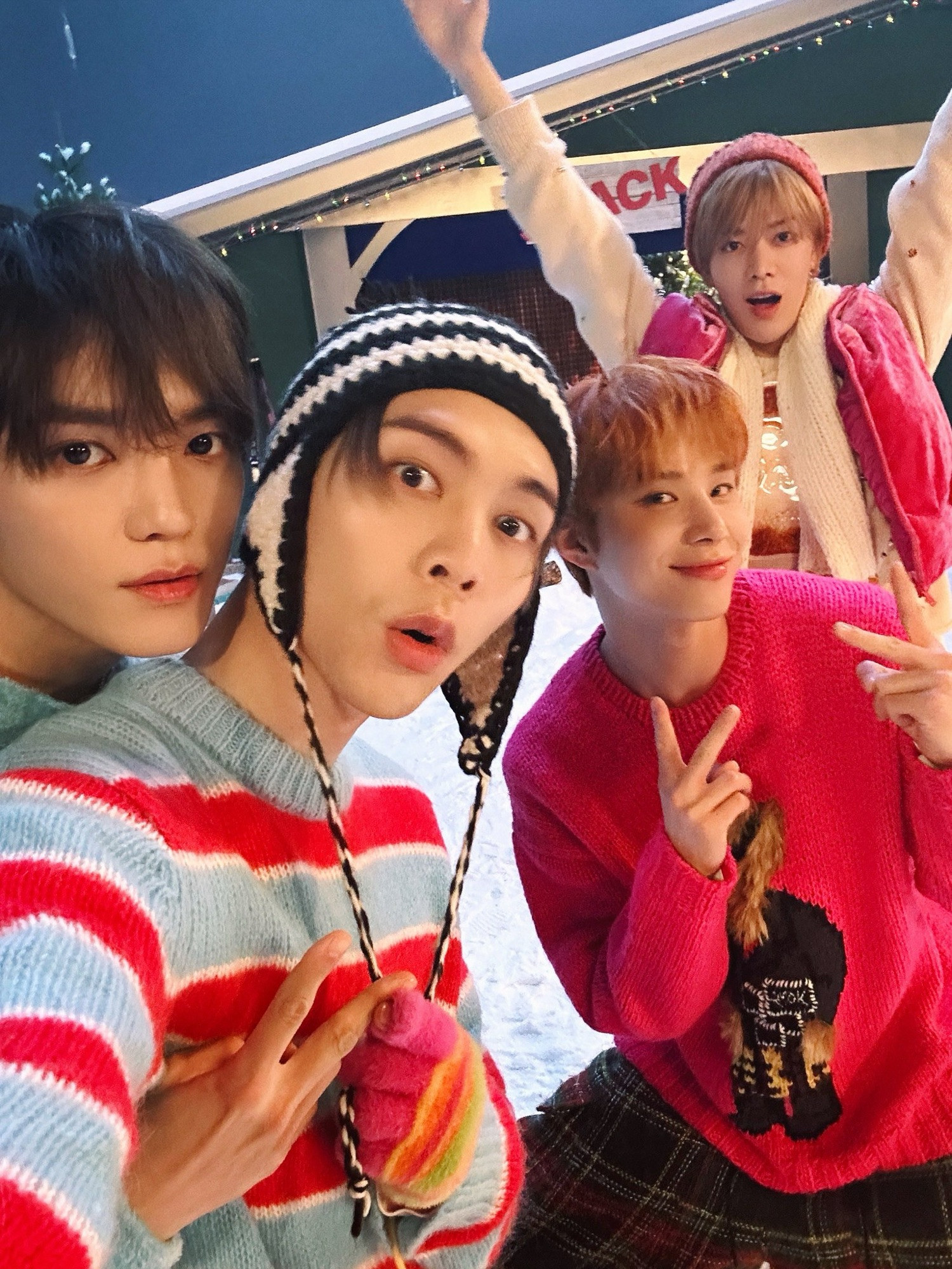 Taeyong, Johnny, Jungwoo, and Yuta on the set of the Be There For Me music video, dressed in winter apparel. Taeyong is giving Johnny a back hug. Johnny is taking the picture and using his free hand to pull down the long dangling strings on his black and white horizontal striped knit winter hat. Jungwoo is wearing a pink sweater and throwing up two peace signs. Yuta is standing behind Jungwoo, wearing a pink puffy vest and knit hat, with both arms in the air.