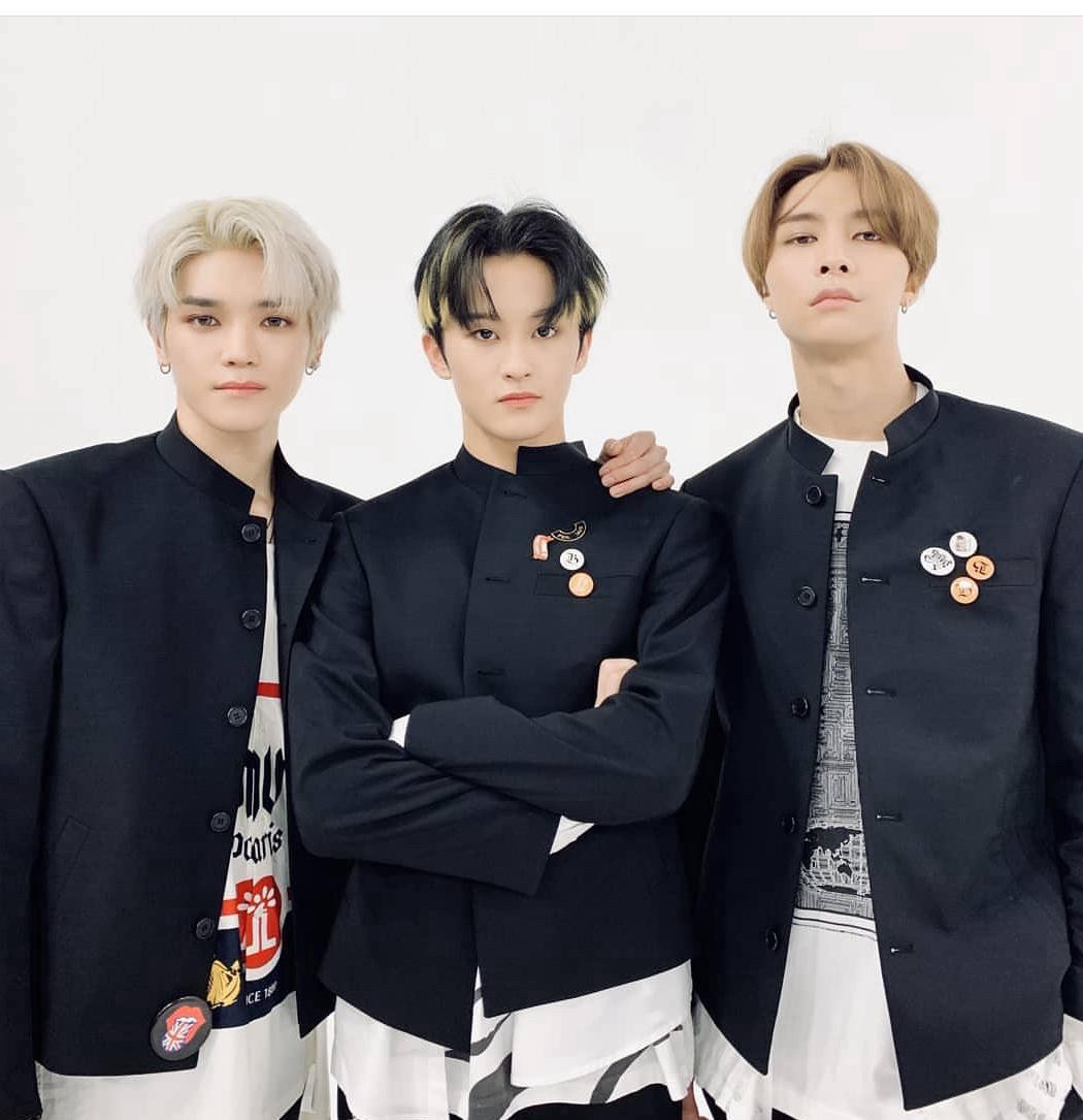 Taeyong, Mark, and Johnny wearing navy blue blazer stage outfits during Kick It era. Taeyong has his arm around Mark’s shoulders. Mark has his arms crossed. Johnny has his hands in his pockets.