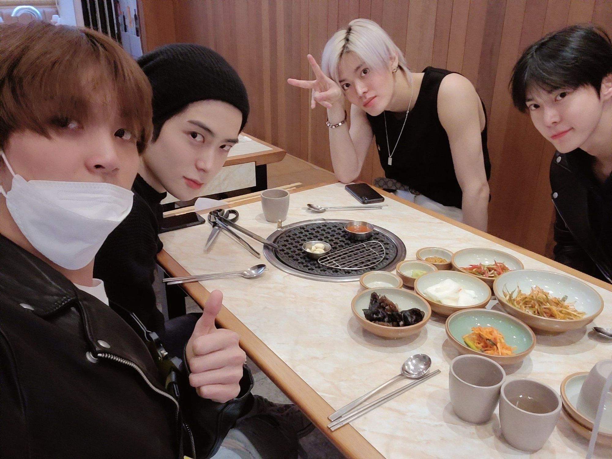 A selfie taken by Haechan of him, Jaehyun, Doyoung, and Yuta sitting at a restaurant table. There is a grill in the middle of the table and plates of side dishes, utensils, and cups of water on the table v