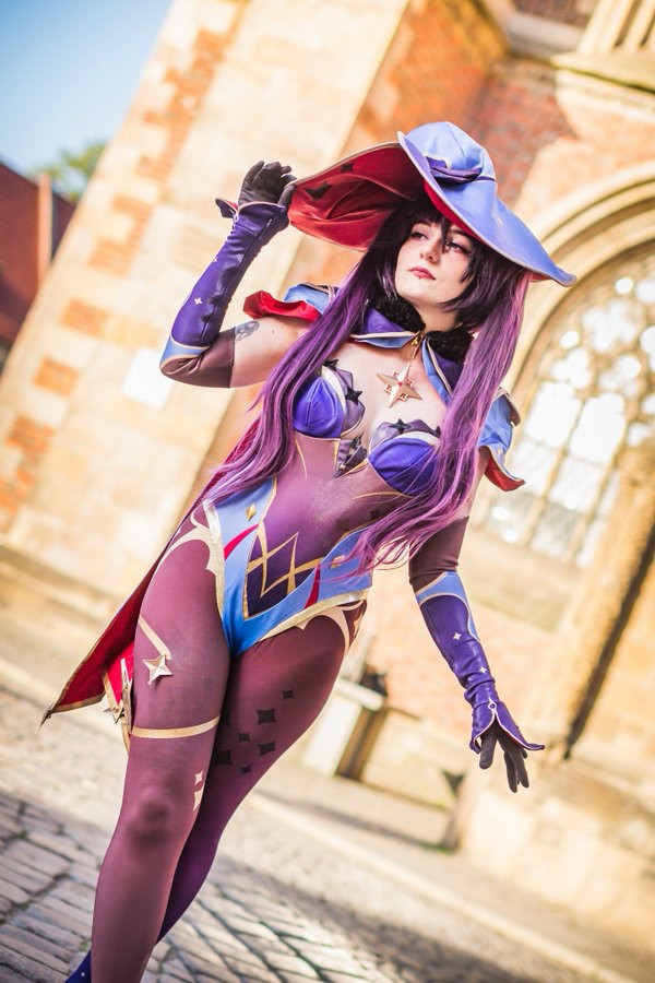 Photo of Nell in a Mona cosplay, consisting of a low-cut blue-purple backless leotard with a detached blue-purple sleeves on their forearms and black gloves, along with a short blue-purple cloak, buttoned with a gold compass rose ornament hanging above their chest. The costume also includes black translucent tights decorated with golden ornaments and a large blue-purple witch hat, adorned with gold compass rose decals and ornaments.
Theyre holding the edge of their hat with their right hand and are looking to the right of the picture.