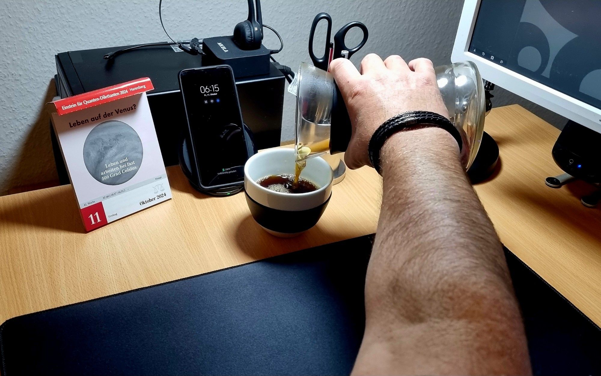 Schreibtischszene - ich schenke mir einen Kaffee ein, im Hintergrund ein Kalender und ein Smartphone mit Ladestation. Das Kalenderblatt fragt, ob es Leben auf der Venus geben könnte. 