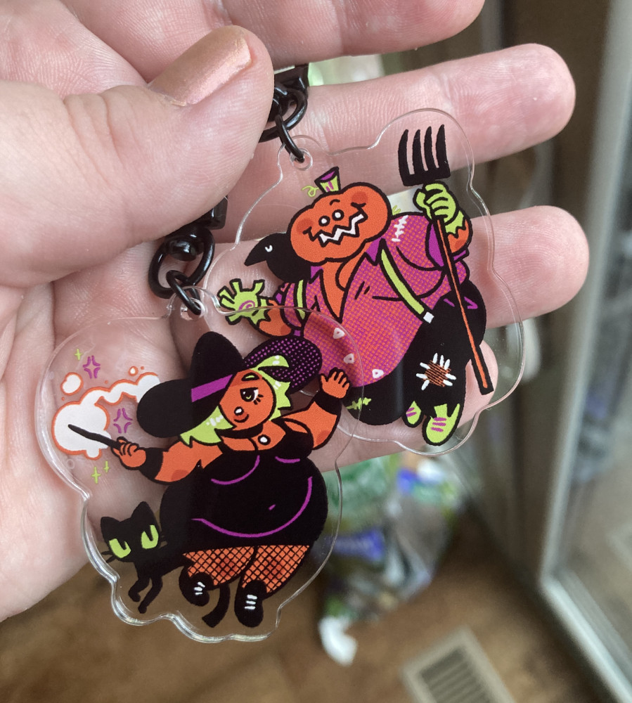 a photo of two new acrylic charms from Emmbunn.bigcartel.com: one is a witch in a little black dress, waving a wand and accompanied by a small black cat. The other is a pumpkin-headed farmer, with a crow companion and a pitchfork.