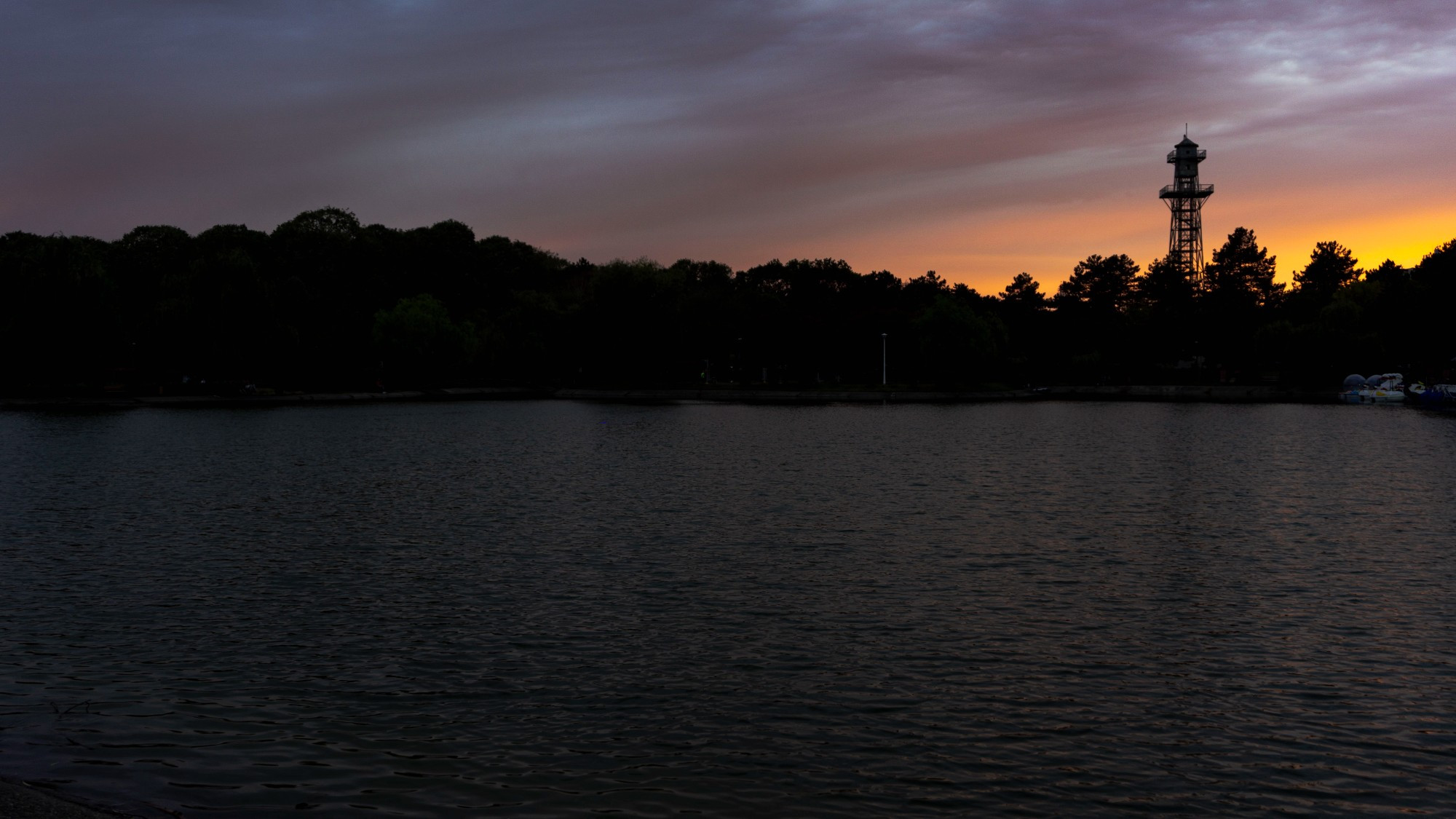 A high contrast picture of a sunset where the sun sets in the right side of the picture. A tower is visible in the right side of the photo and most of the lower part is a slightly agitated lake. The middle is the silhouette of trees and upwards is the cloudy sky.