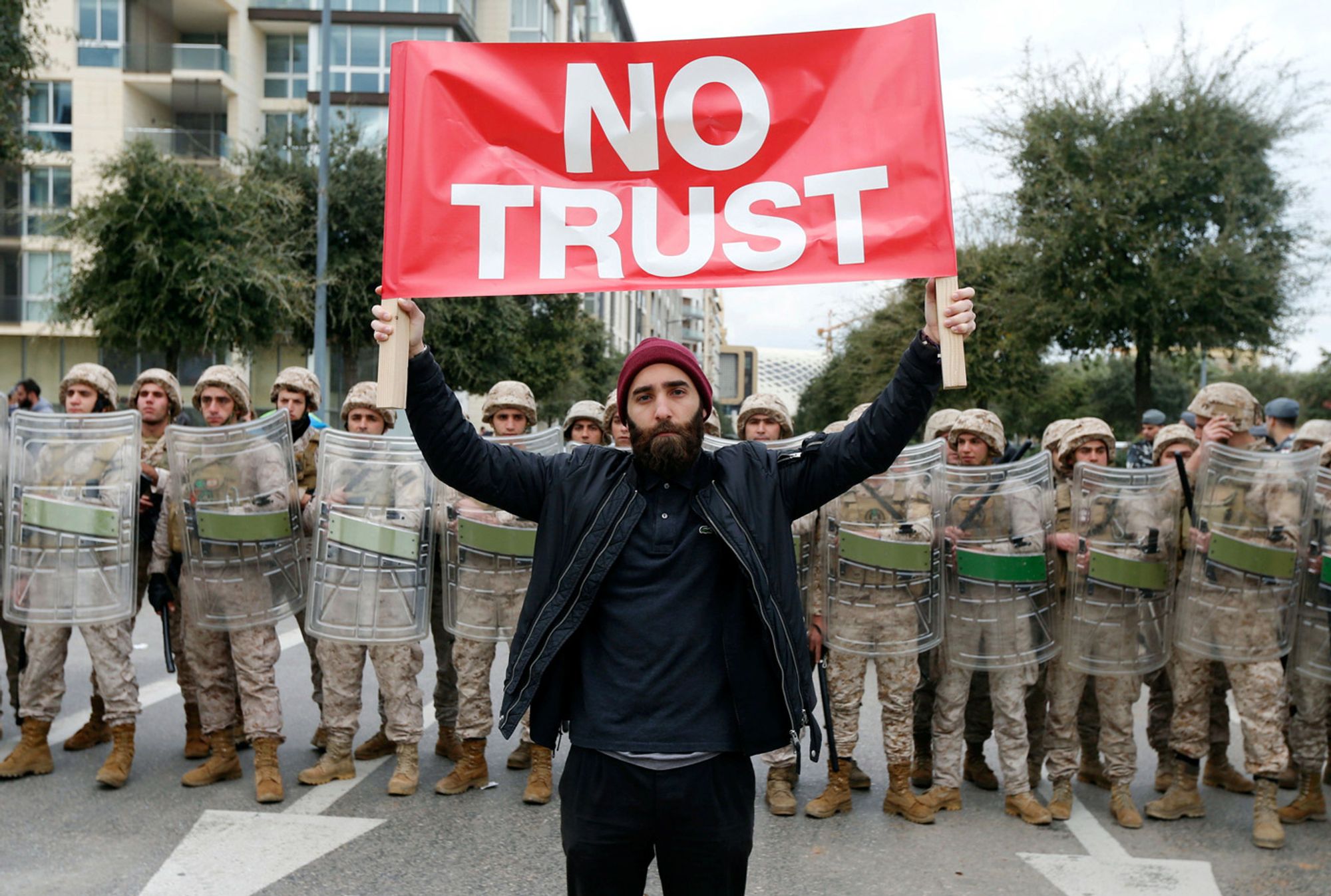 A demonstrator in the wave of protest that began in Lebanon in 2019 stands in front of a line of militarized officers in armor, holding up a banner reading "No trust."