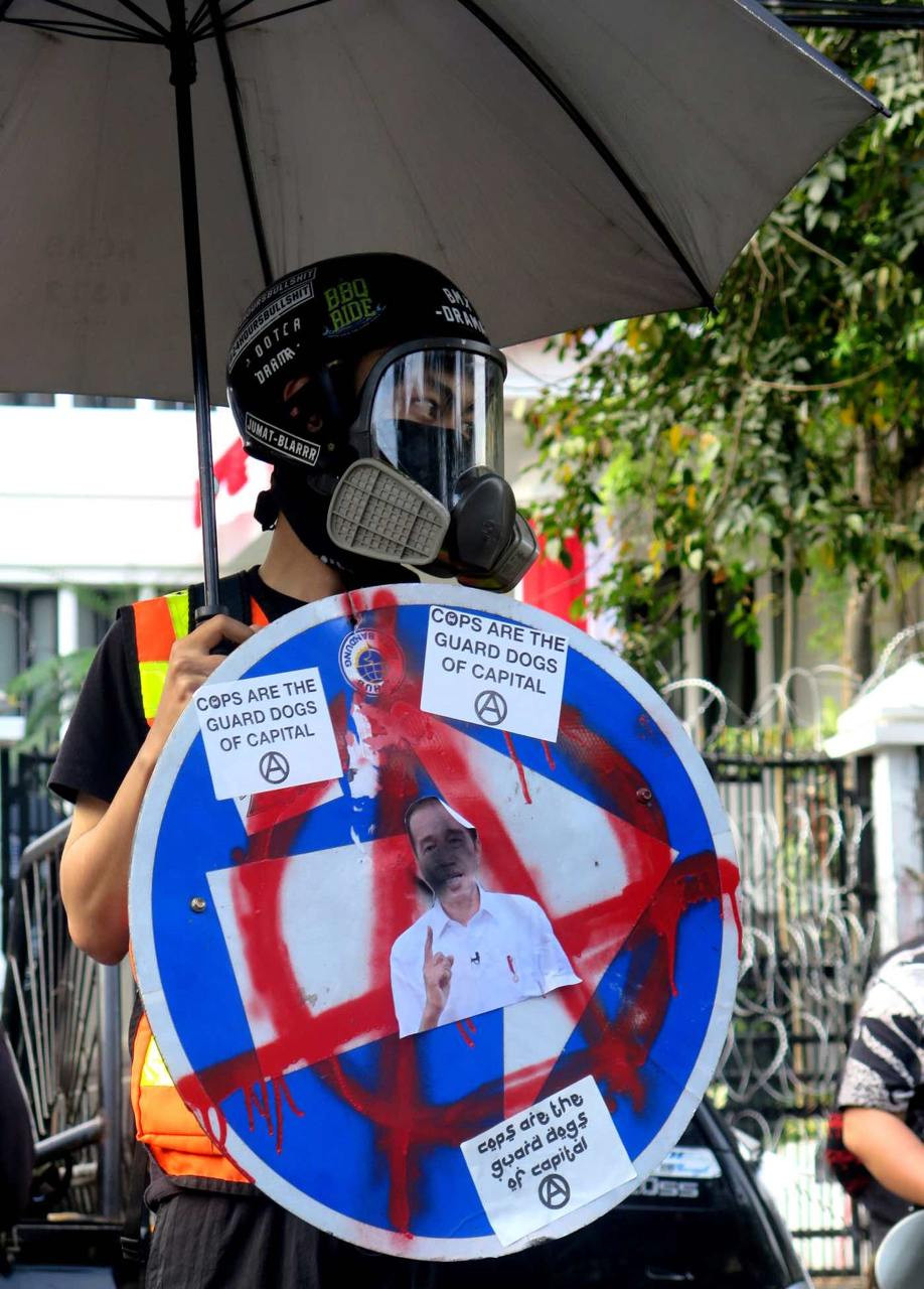 A protester wearing a gas mask wields an umbrella and a street sign with a circle-A (the universal symbol for opposition to all forms of domination) spray-painted on it at a demonstration in Bandung on August 28, 2024.