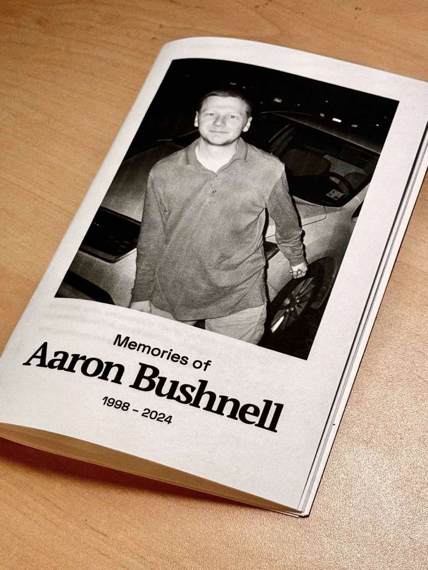 A photograph of the cover of a zine named "Memories of Aaron Bushnell 1998-2024." The cover shows a black-and-white picture of Aaron wearing a long-sleeved shirt—a red shirt that his friends remember him often wearing—standing in front of a car.