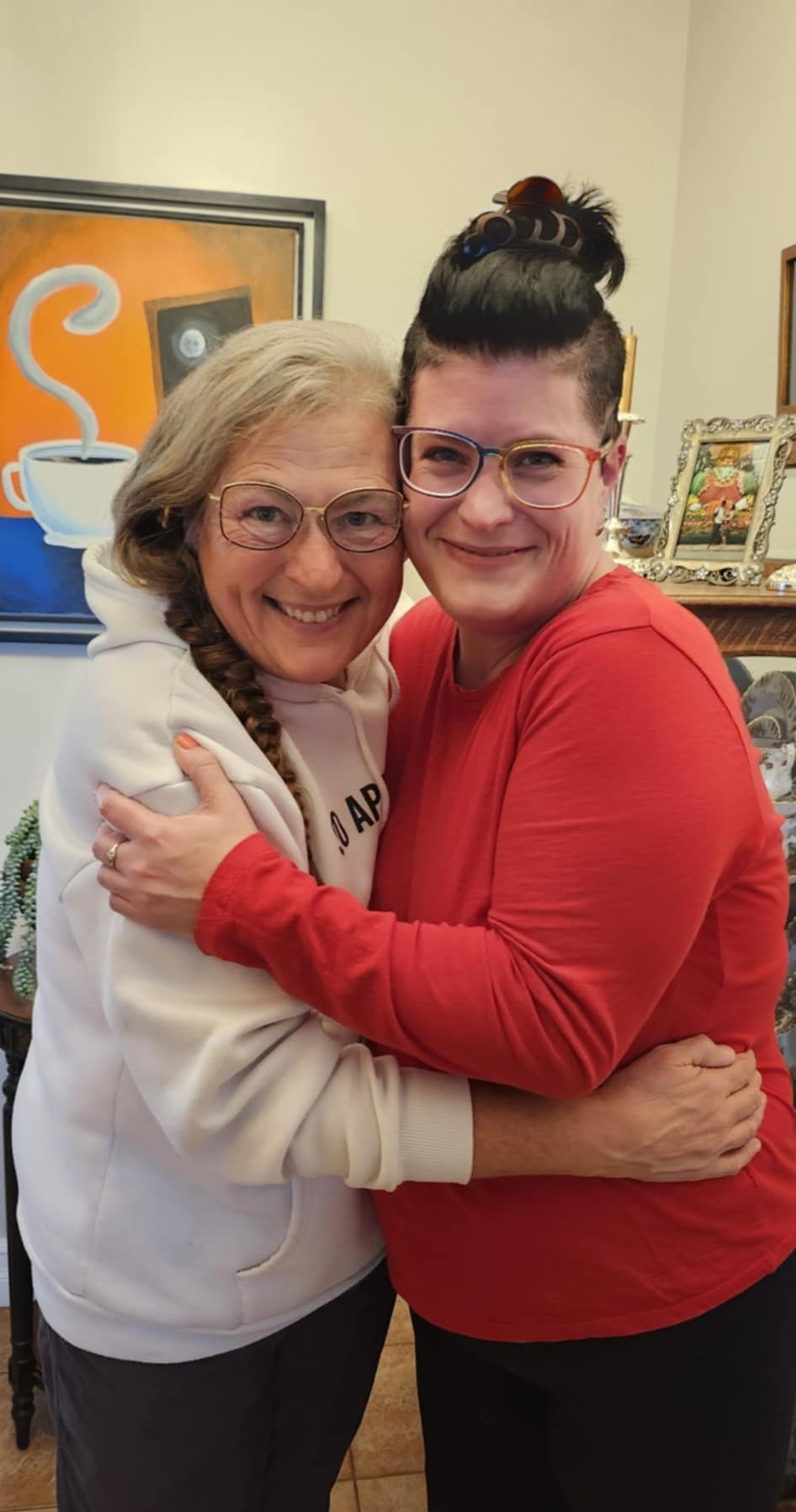 A woman with a braid in a white hoodie hugging Kelly in a red shirt. Both women are smiling. 