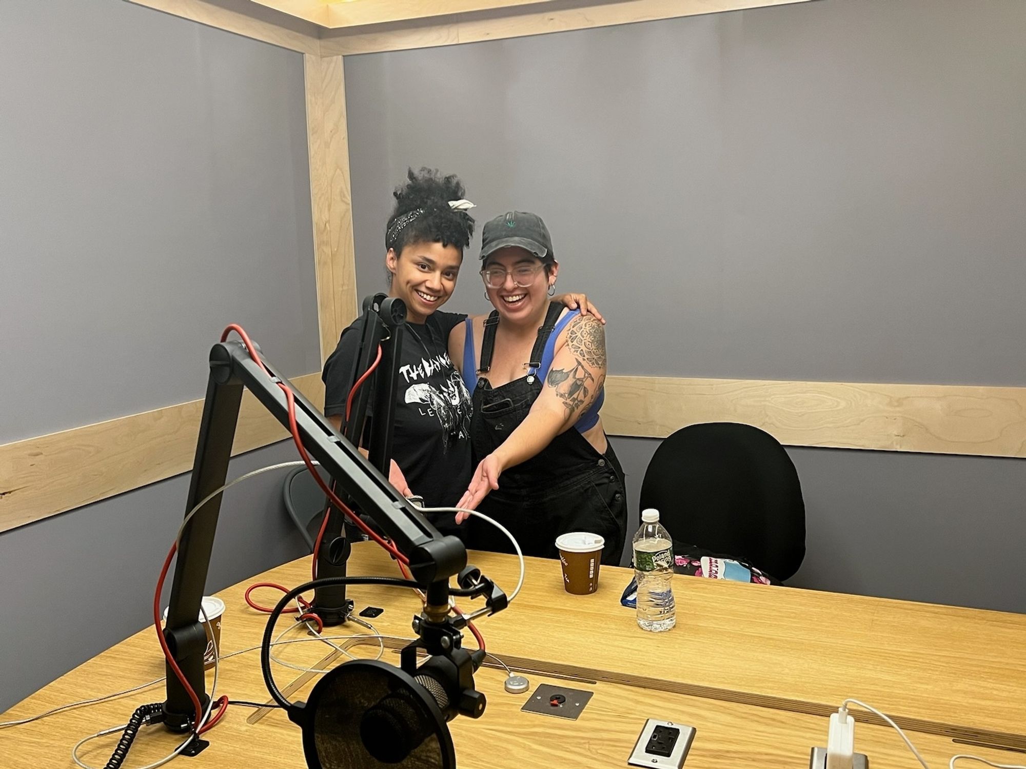 Two dykes standing in a recording studio smiling and pointing at a microphone