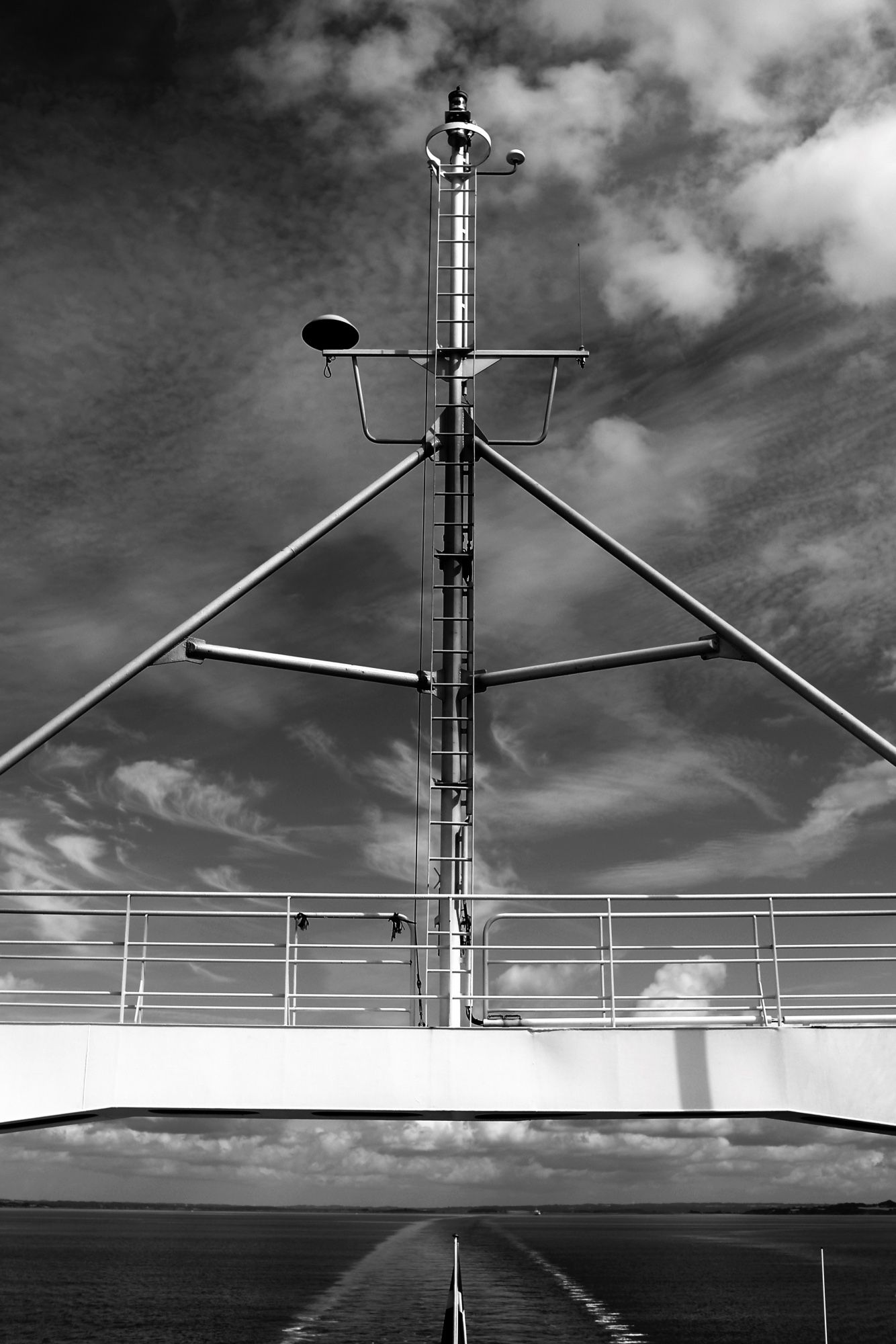 Black and white shot from the deck of a RoRo ferry with a bridge and a signal mast over to the coast where the journey began – with an interesting skyscape over the calm sea.