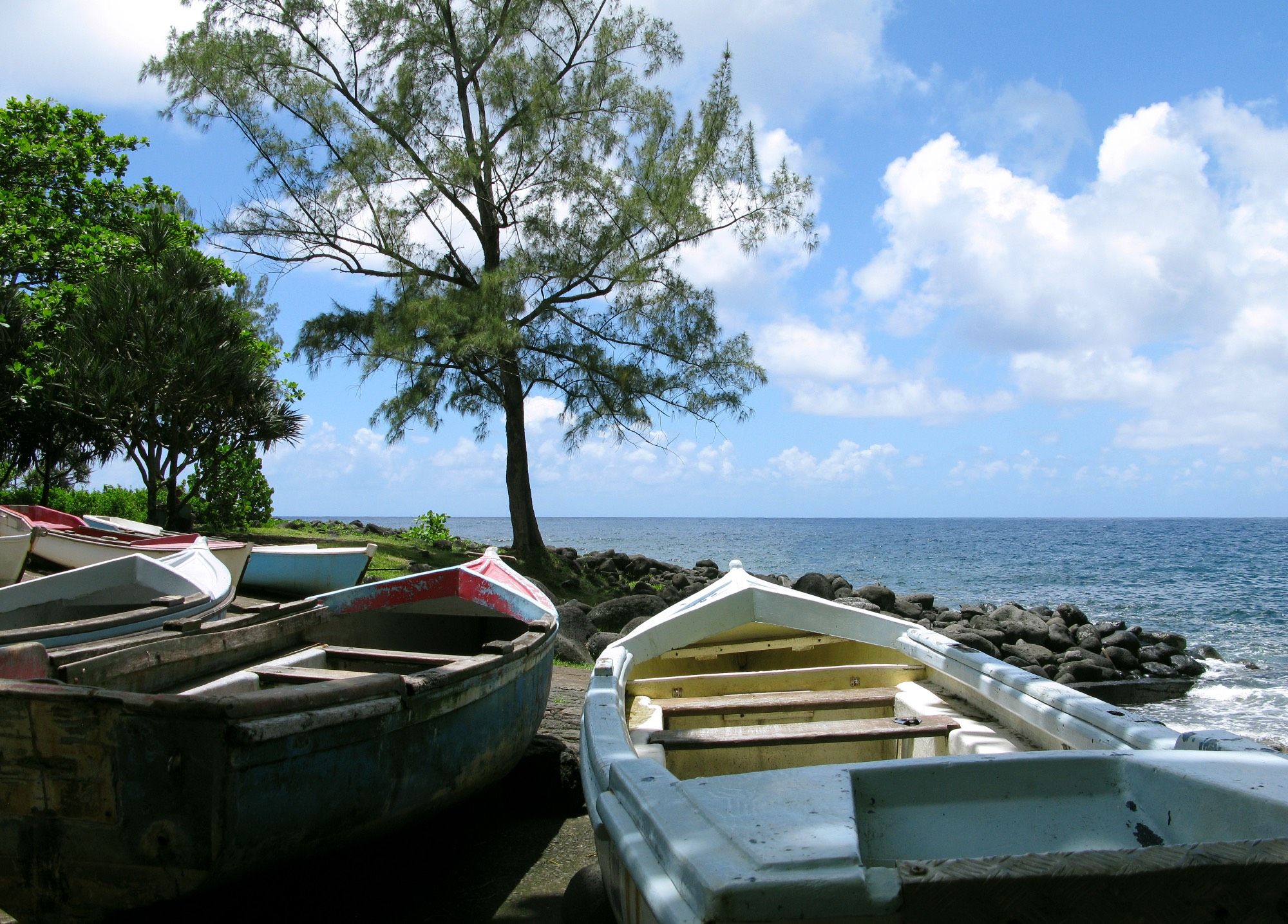 Boote im Halbschatten an Land in einem kleinen, verträumten Fischerhafen beim Restaurant L'Anse des Cascades am Rand von Sainte-Rose auf der französischen Übersee-Insel La Reunion. Im Bild blauer Himmel mit ein paar weißen Wolken, zwei Bäume, der weite, türkisblaue Indische Ozean sowie ein Stückchen Lavaküste.

© Torsten Grieger