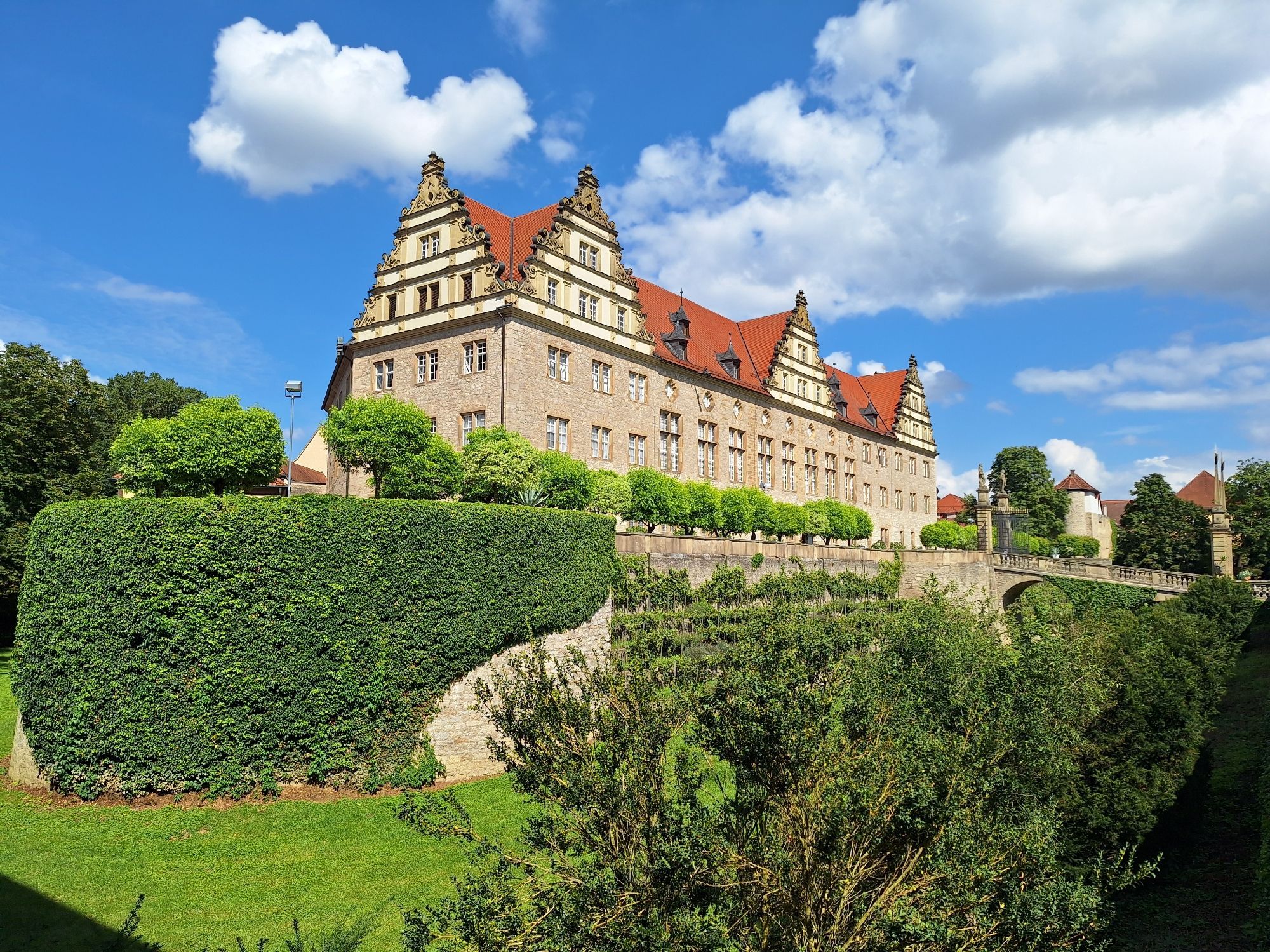 Großes Schloss mit drei Giebeln auf einem Bergfried seitlich fotografiert. Umgeben von einem Graben, an dessen Hängen Reben stehen, Efeu wächst und unten eine Wiese ist.