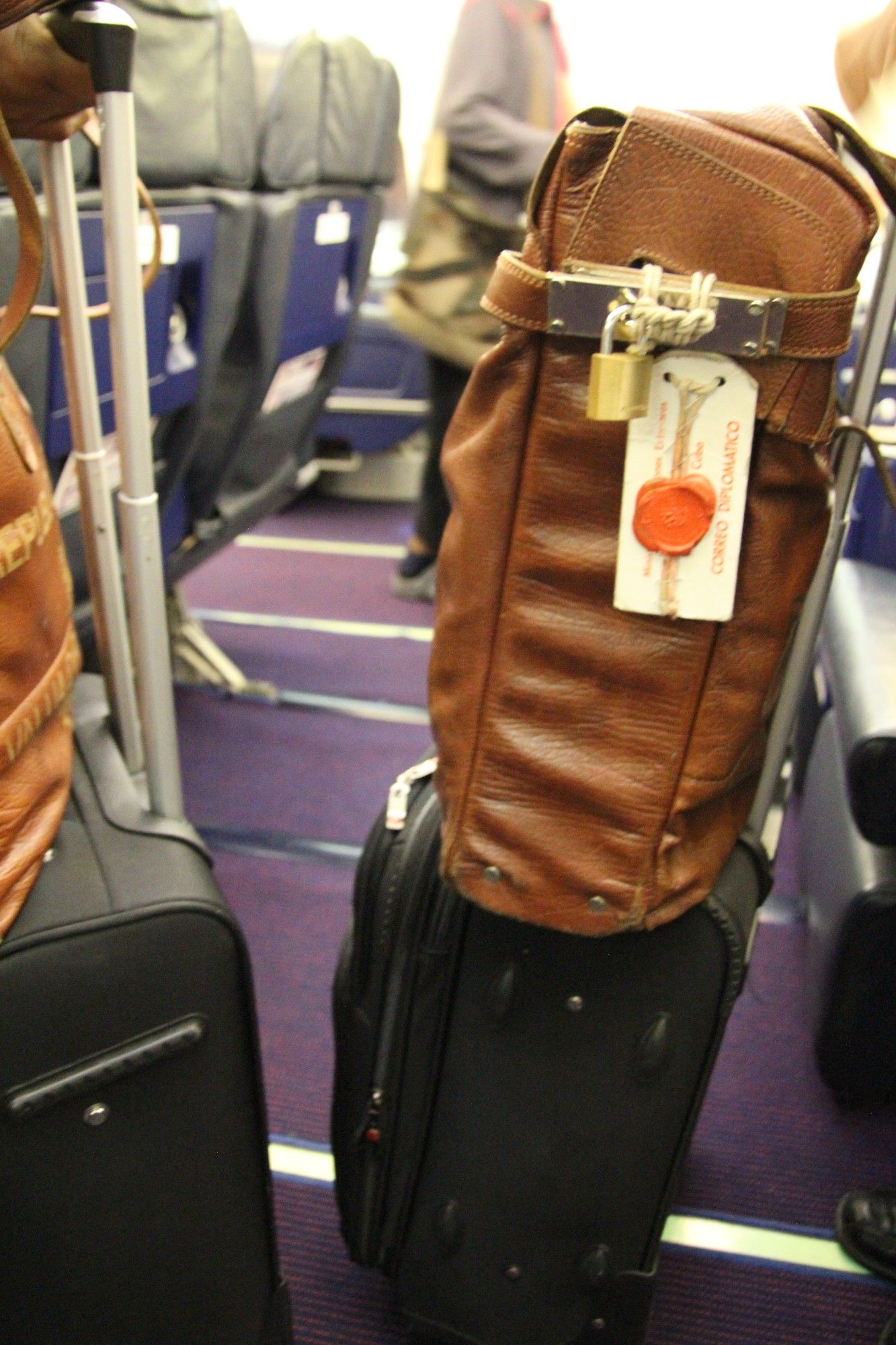 brown leather back with a padlock and tag "correo politico" on top of a black roller suitcase.