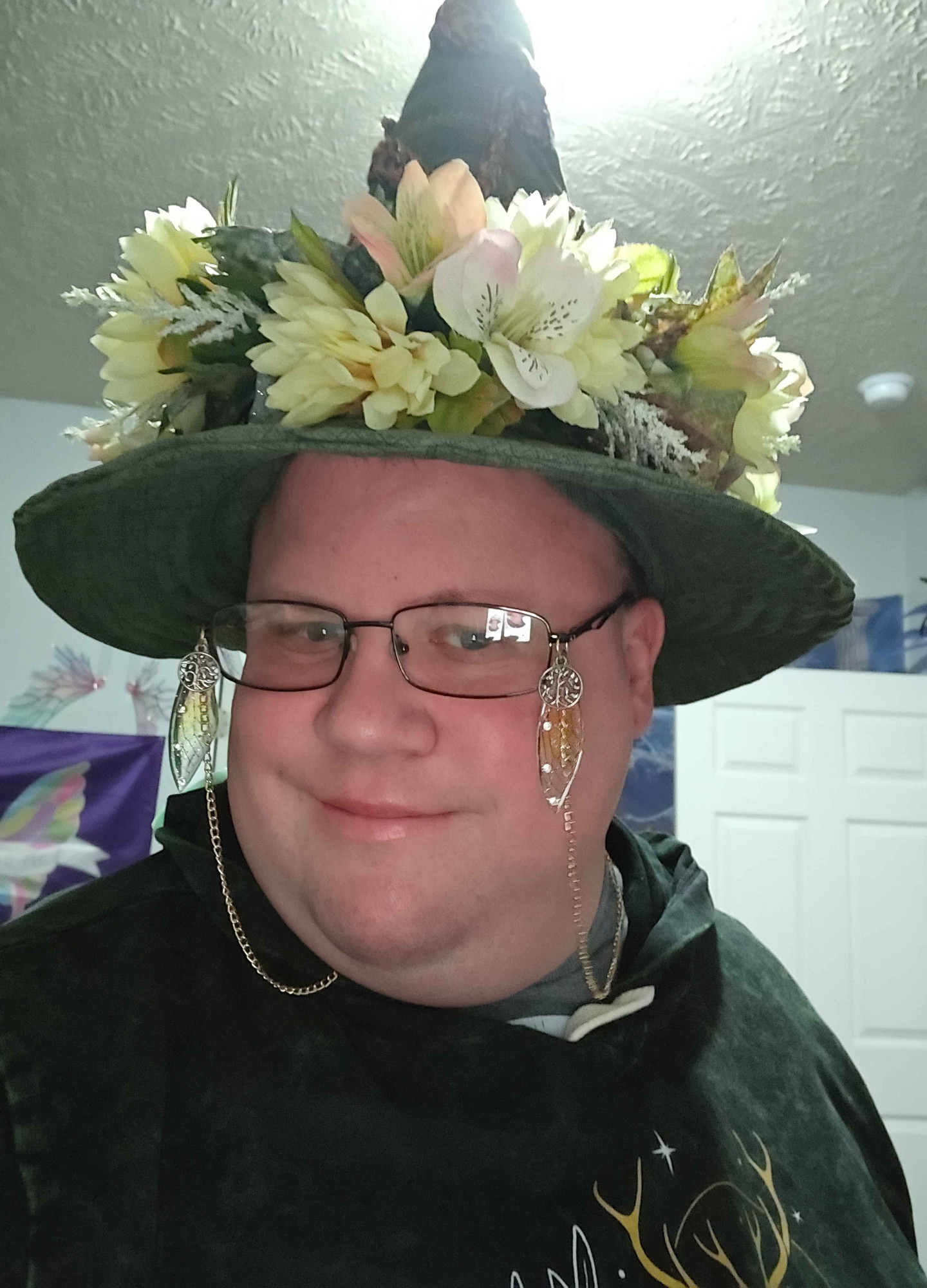 Close-up selfie, showing a gold glasses chain. Hanging off the temples of the chain are small silver tree of life medallions, and faerie wings in yellow hues.