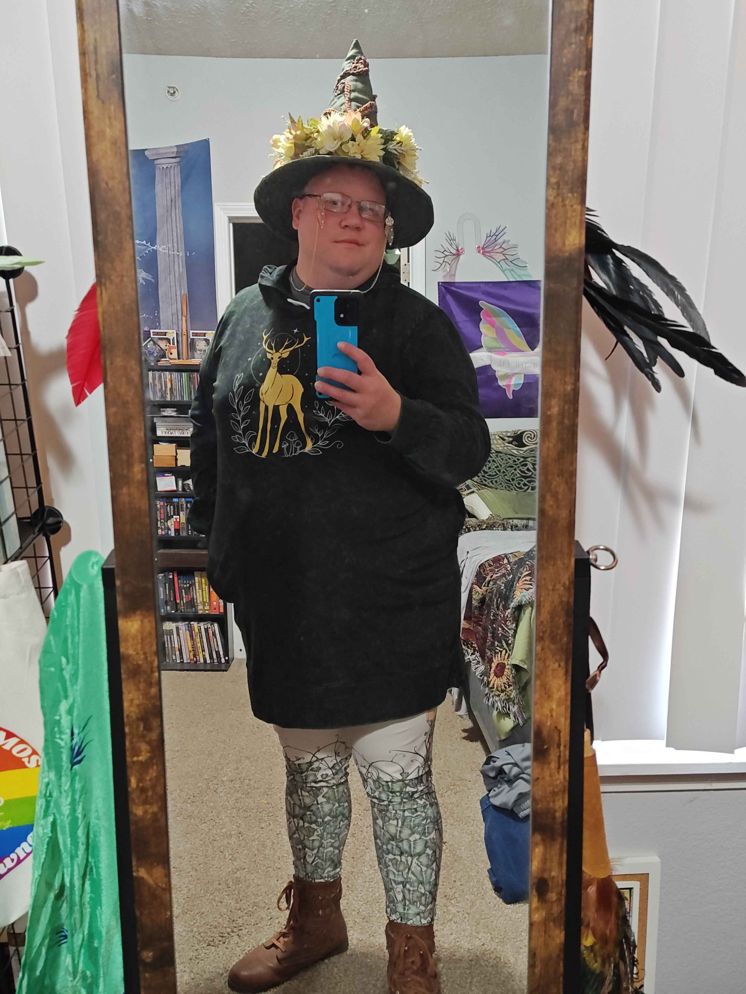 Mirror selfie of myself. Wearing a green witch hat with brown stitching, and a white flower crown around the base of it, a sweater dress in dark green with a deer across the chest in golden ink, cream-colored leggings with twisting vine print around the calves, and light brown ankle boots.