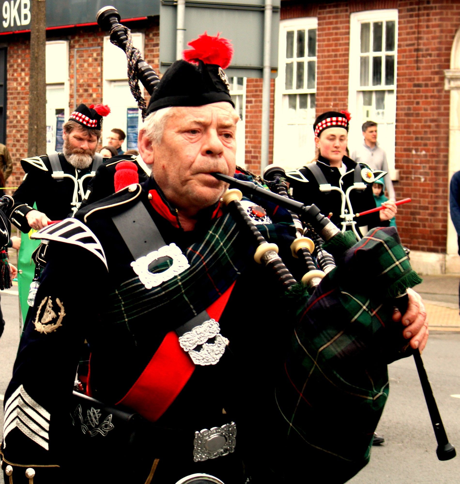 An older man playing bagpipes, marching in a parade.