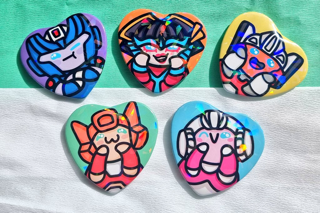 6 heart shaped badges, with a glittery holo effect. They depict from left to right: Chromia, Elita-one, Windblade, Arcee and Minerva from Transformers. 
They're cute, colourful with bold outlines and vivid colours.