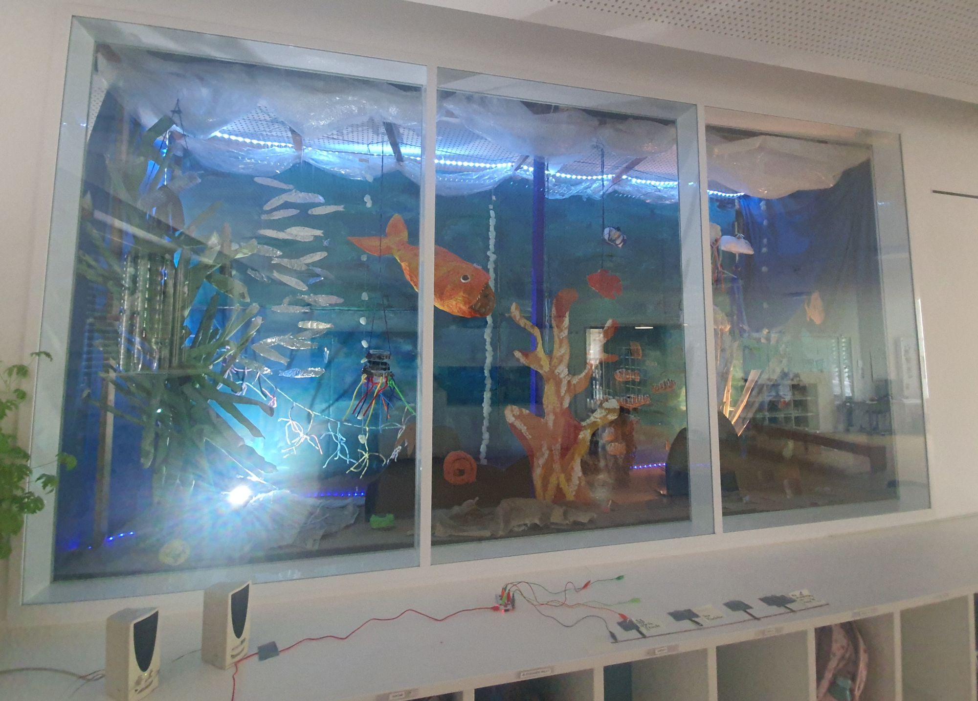 A window between 2 rooms, transformed into a huge aquarium with cardboard and papier-mâché. Corals can be seen in the foreground and a large orange anglerfish swims in the middle. There are loudspeakers on a cupboard in front of the window. A makeymakey board is wired so that you can call up information on individual objects in the aquarium.