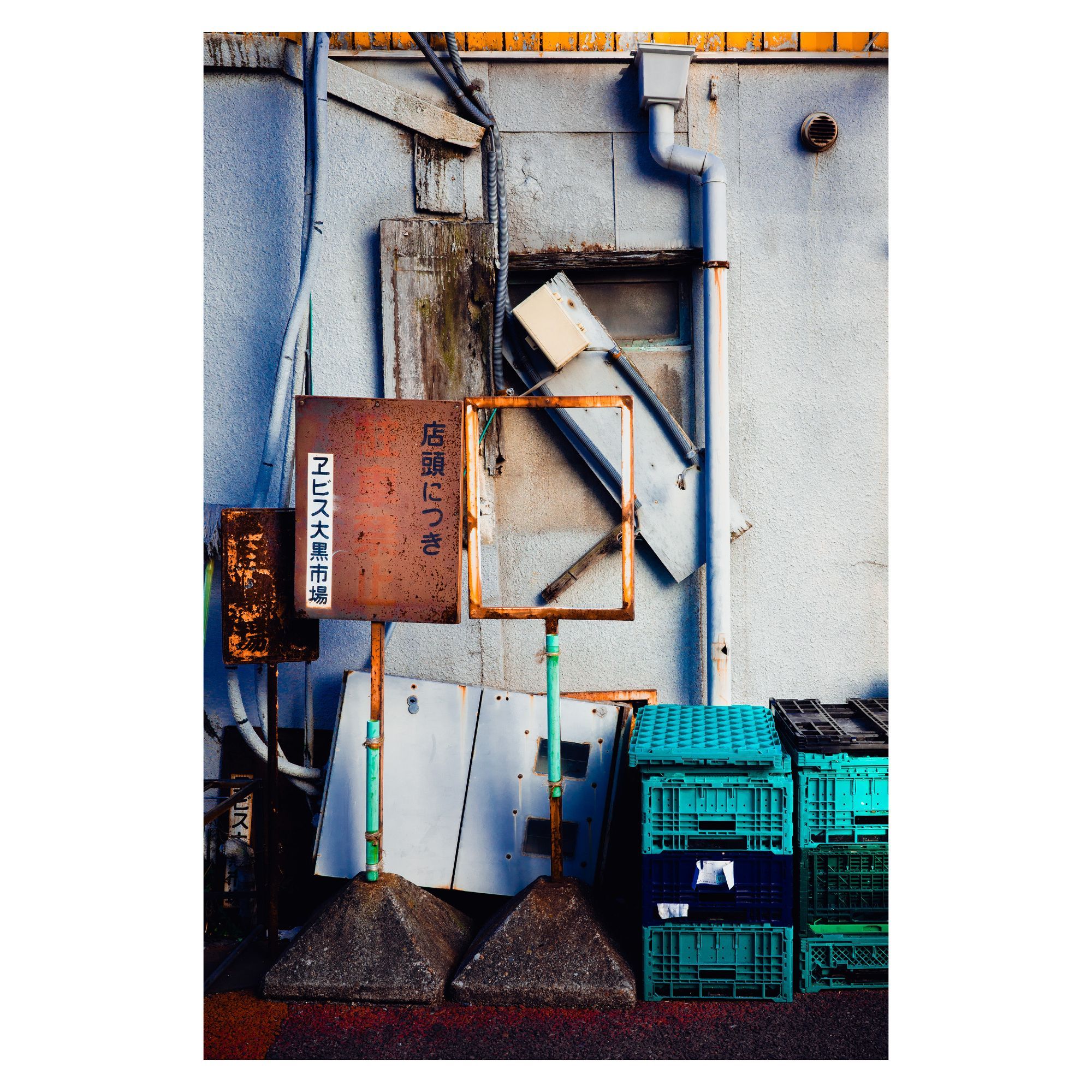 The photo captures a cluttered section of a wall, where rusted metal signs, broken panels, and a stack of plastic crates are haphazardly placed. The rusted signs, one of which reads “店頭につき” (related to a store or shopfront), are weathered and leaning against the wall. Various pipes and vents snake across the wall in the background, while mismatched panels and crates complete the scene. The colors are a mix of muted tones and bright turquoise from the crates, creating a disordered yet visually intriguing composition.